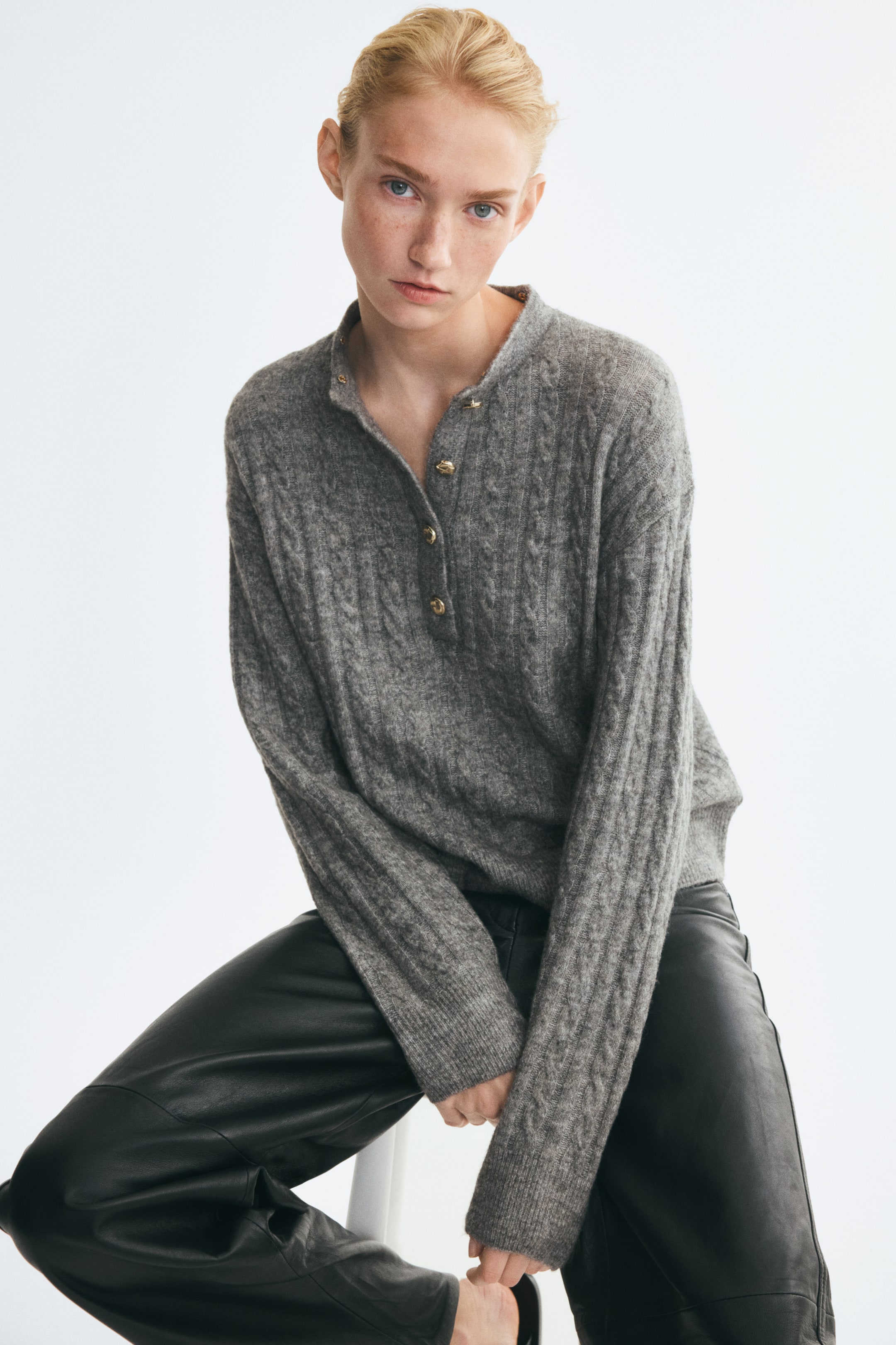 Ver imagen más grande: La modelo lleva puesto un jersey polo gris de punto trenzado, con tapeta de botones y botones en tono dorado, hombros caídos y mangas largas con puños acanalados. El jersey se combina con pantalones de cuero negros.