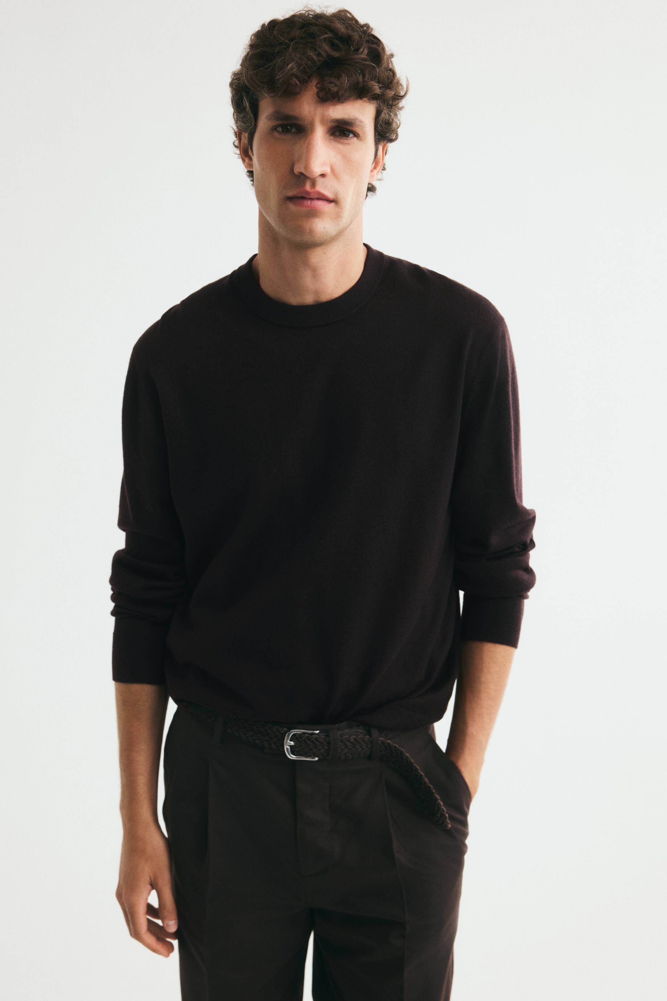 View larger image: The model is wearing a dark brown, fine-knit merino wool jumper with a round, rib-trimmed neckline. The jumper features long sleeves with ribbed cuffs and is styled tucked into dark trousers with a braided belt.