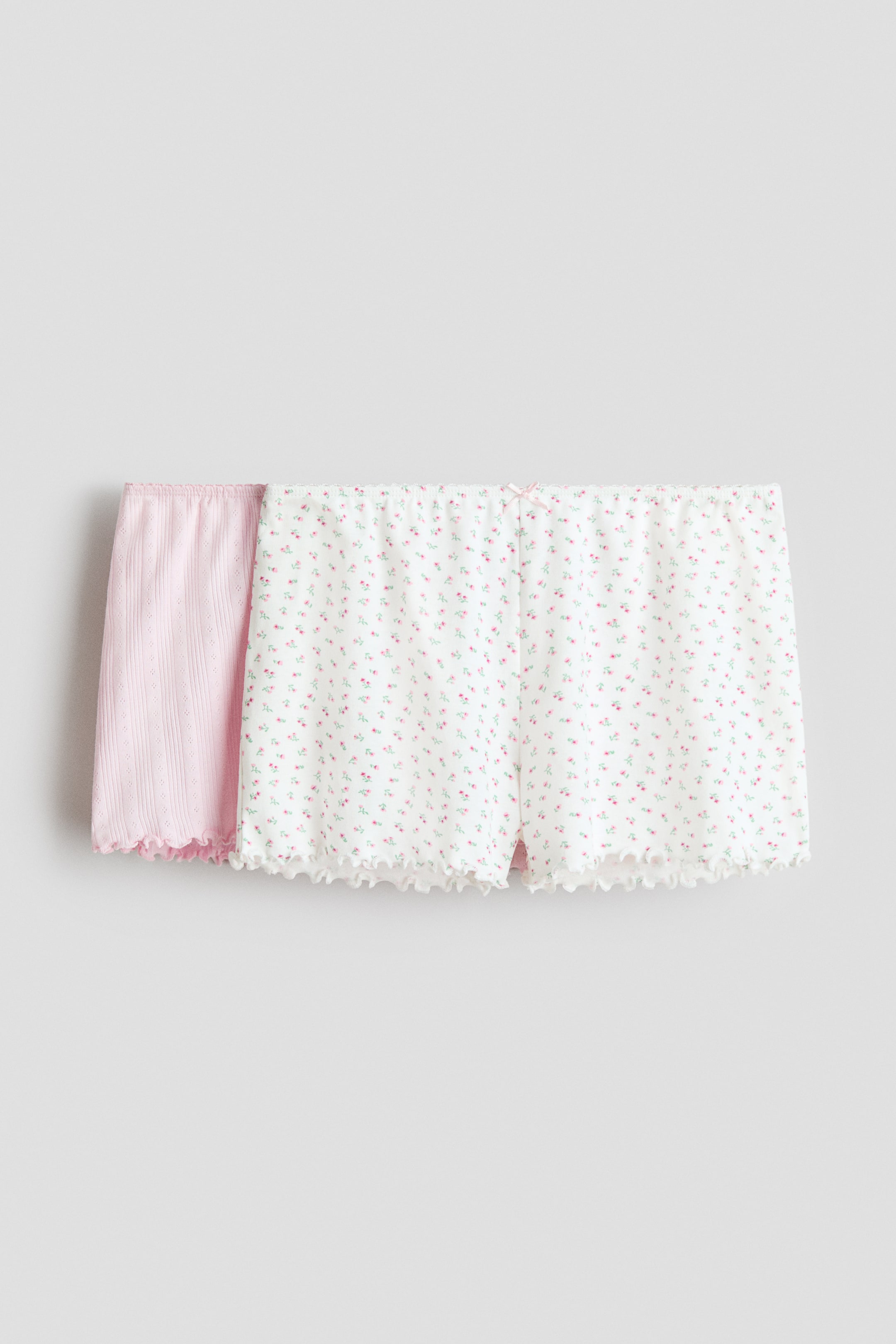 View larger image: Two pairs of shorts are laid flat, with one white pair overlapping a pink pair. The front white shorts feature an allover pink floral print, an elasticated waistband with a small pink bow, and ruffled leg hems, while the pink shorts behind boast a ribbed texture with eyelet details and ruffled leg hems.