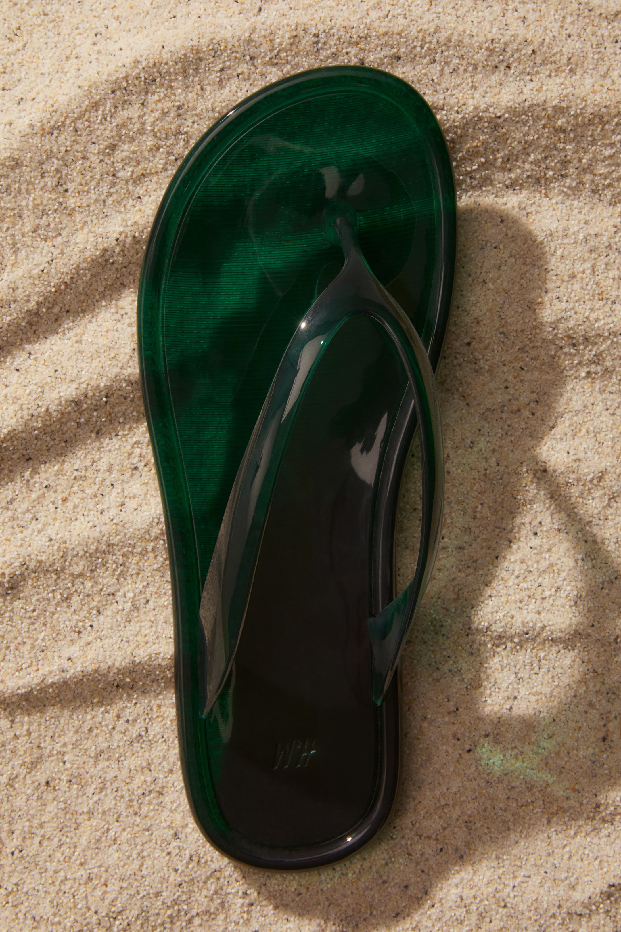 Ver imagen más grande: Una sola chancla de gelatina translúcida de color verde oscuro está colocada plana con la punta hacia arriba. Presenta una suela texturizada y un logotipo visible de 'H&M' grabado en el talón.
