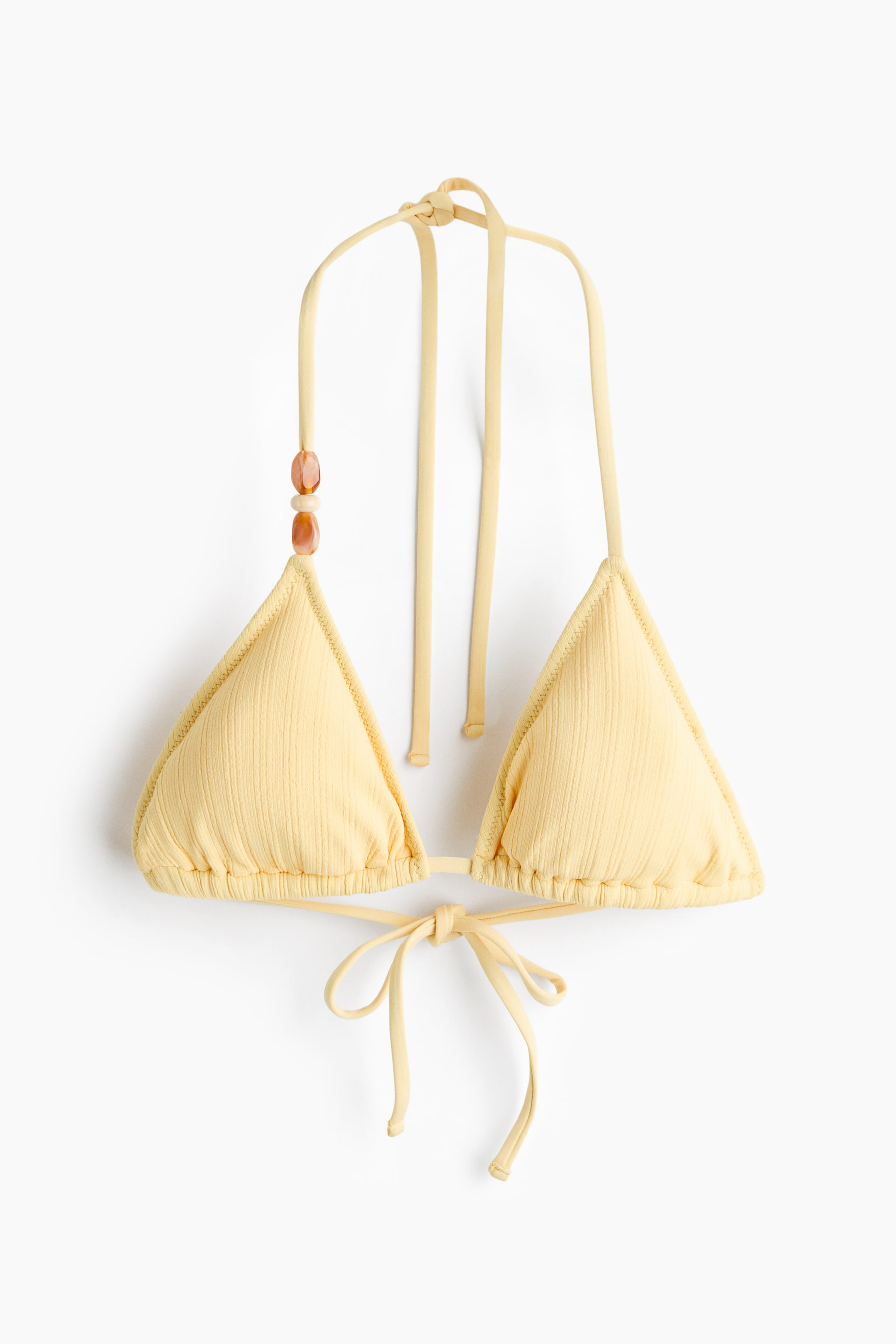 Visa större bild: En trekantsbikiniöverdel med blekgula, ribbade trekantskupor. Knytband formar en rosett i ryggen och en knut i nacken, med tre dekorativa pärlor som pryder ett av nackbanden.