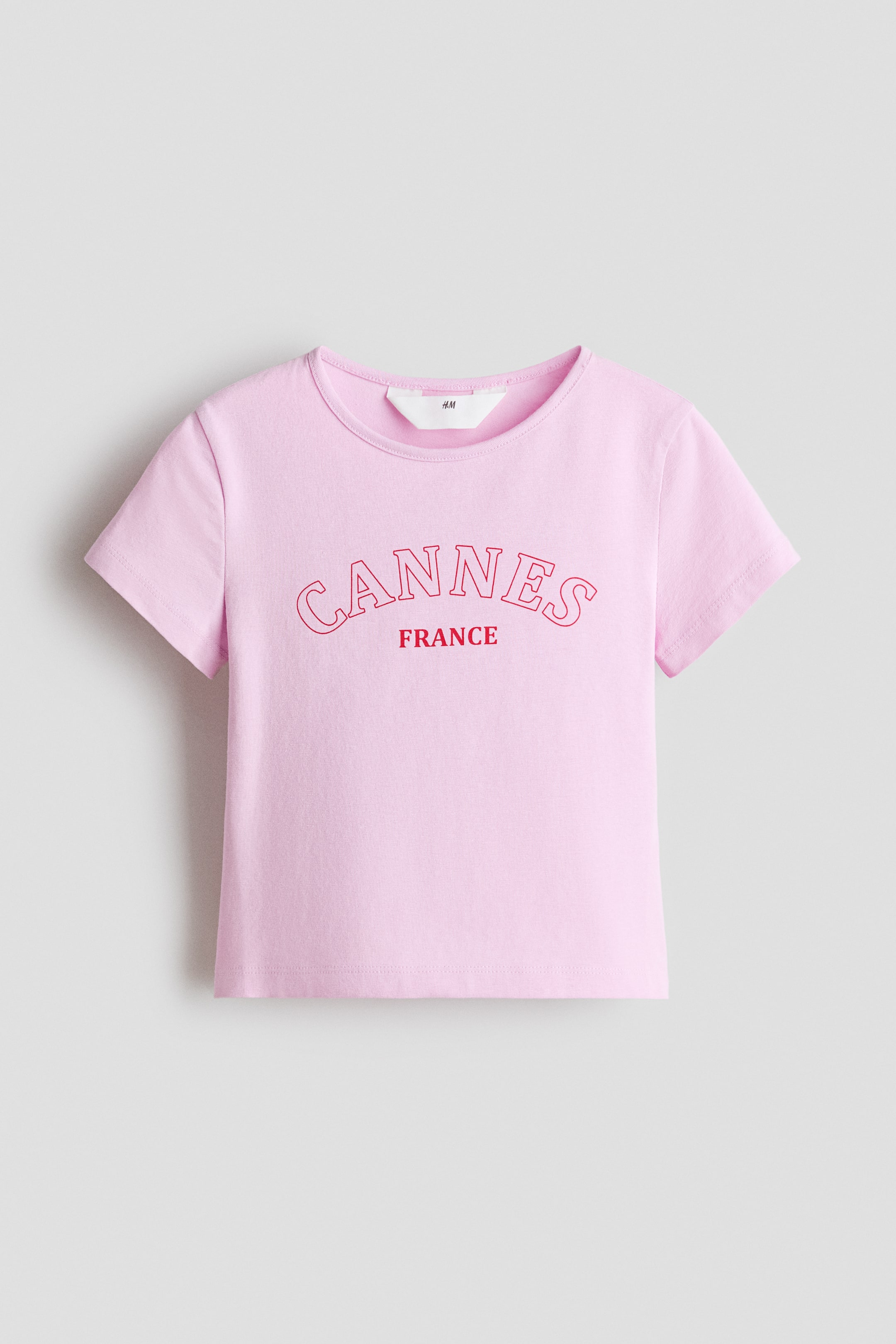 View larger image: A short-sleeved top in soft pink cotton jersey is shown from the front. It features a round neckline and a red print motif on the front reading 'CANNES' above 'FRANCE'. A white H&M label is visible inside the back neckline.
