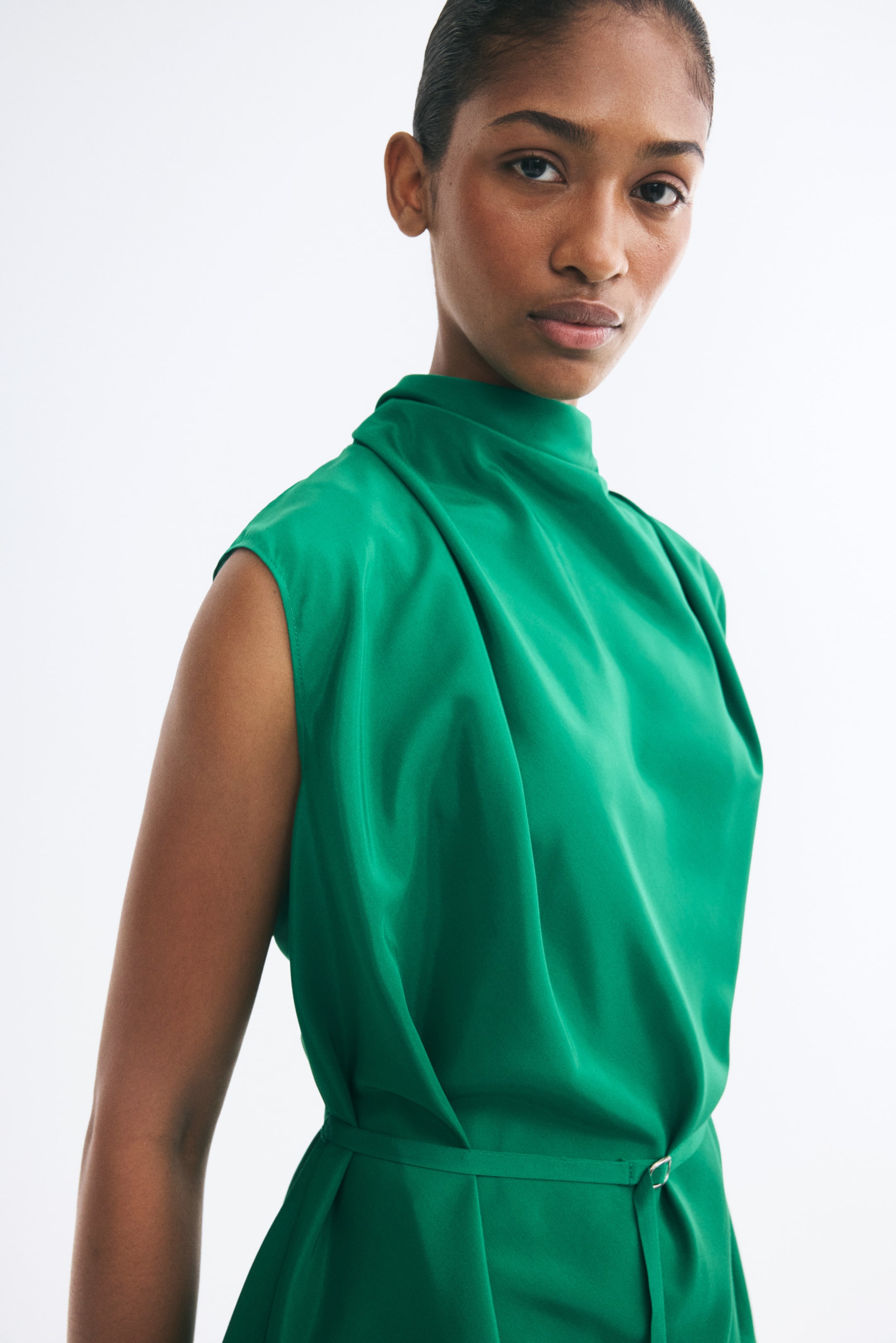 View larger image: A green sleeveless top with a draped cowl neckline and shoulder pleats is worn by the model. The top is secured at the waist with a narrow, adjustable matching belt featuring a metal slide buckle.