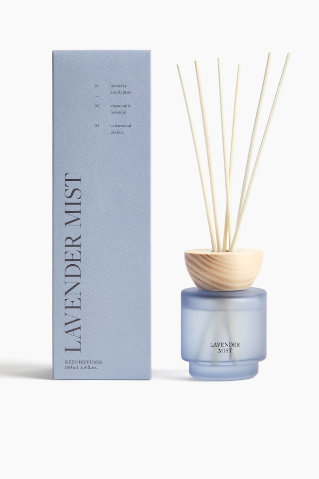 A reed diffuser set features a frosted light blue glass bottle containing scented oil, with a light wooden ring encircling its opening. Several light wooden reeds are protruding from the bottle. To the left, a matching light blue rectangular box showcases product information, including "LAVENDER MIST" and "100 ml 3.4 fl. oz.". - 1