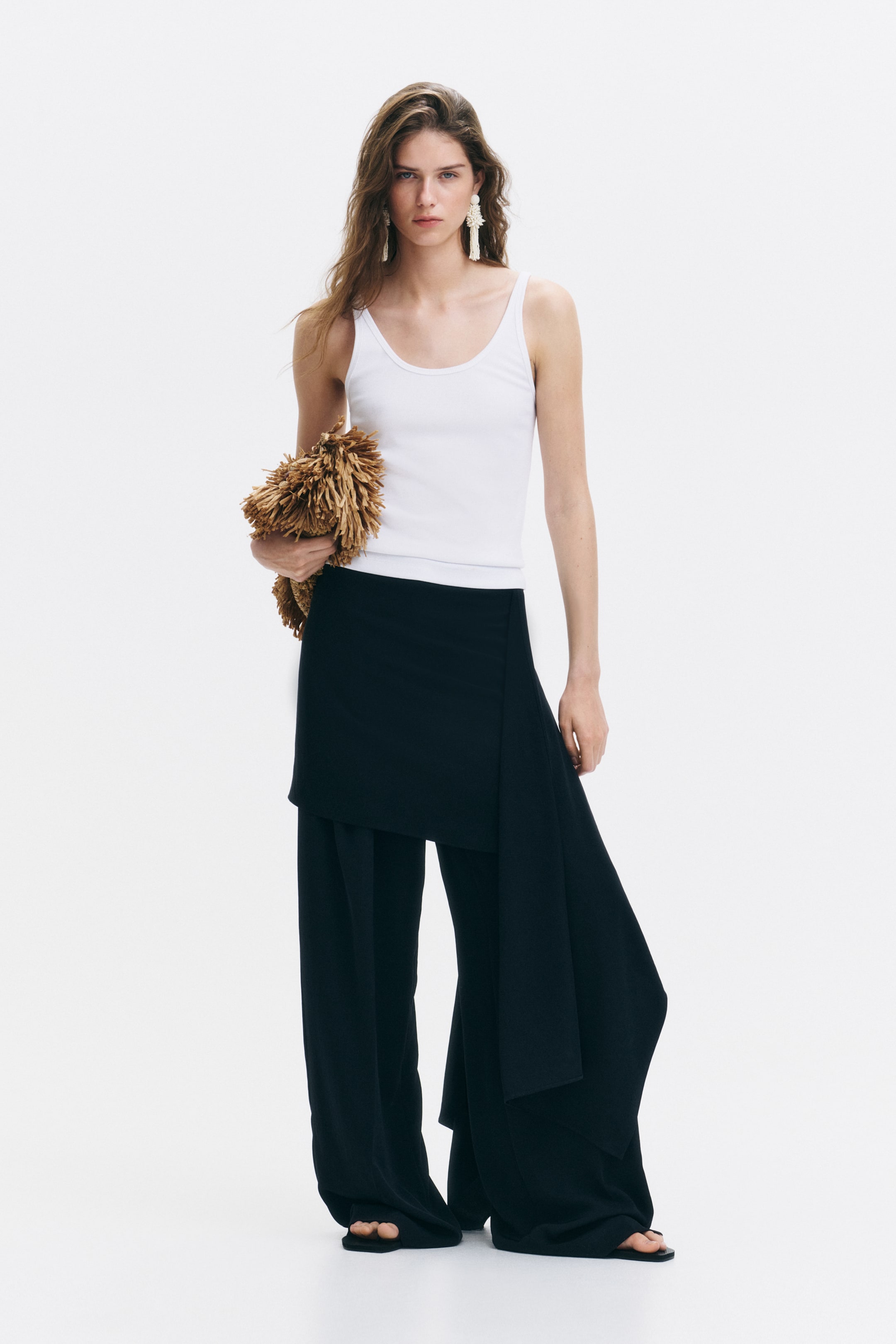 View larger image: A fitted white ribbed jersey vest top with a scoop neckline at the front and back is being worn by the model, tucked into black trousers.