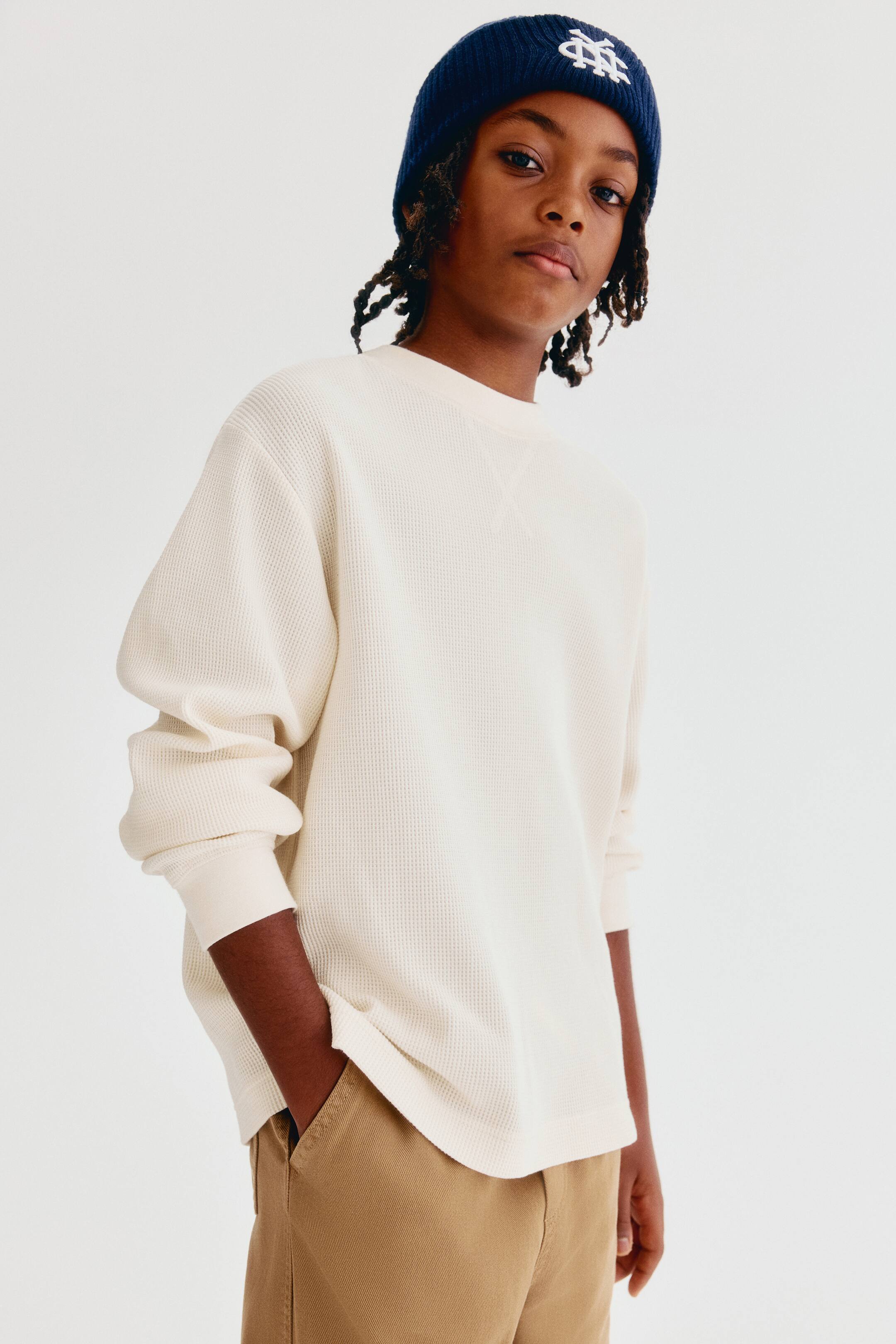 View larger image: A cream-coloured, loose-fit tee in a textured waffle-knit jersey is shown from the front, featuring a round neckline, dropped shoulders, and long sleeves with ribbed cuffs. The soft, textured top drapes loosely, with one side of the hem slightly raised where it meets the pocket of khaki trousers.