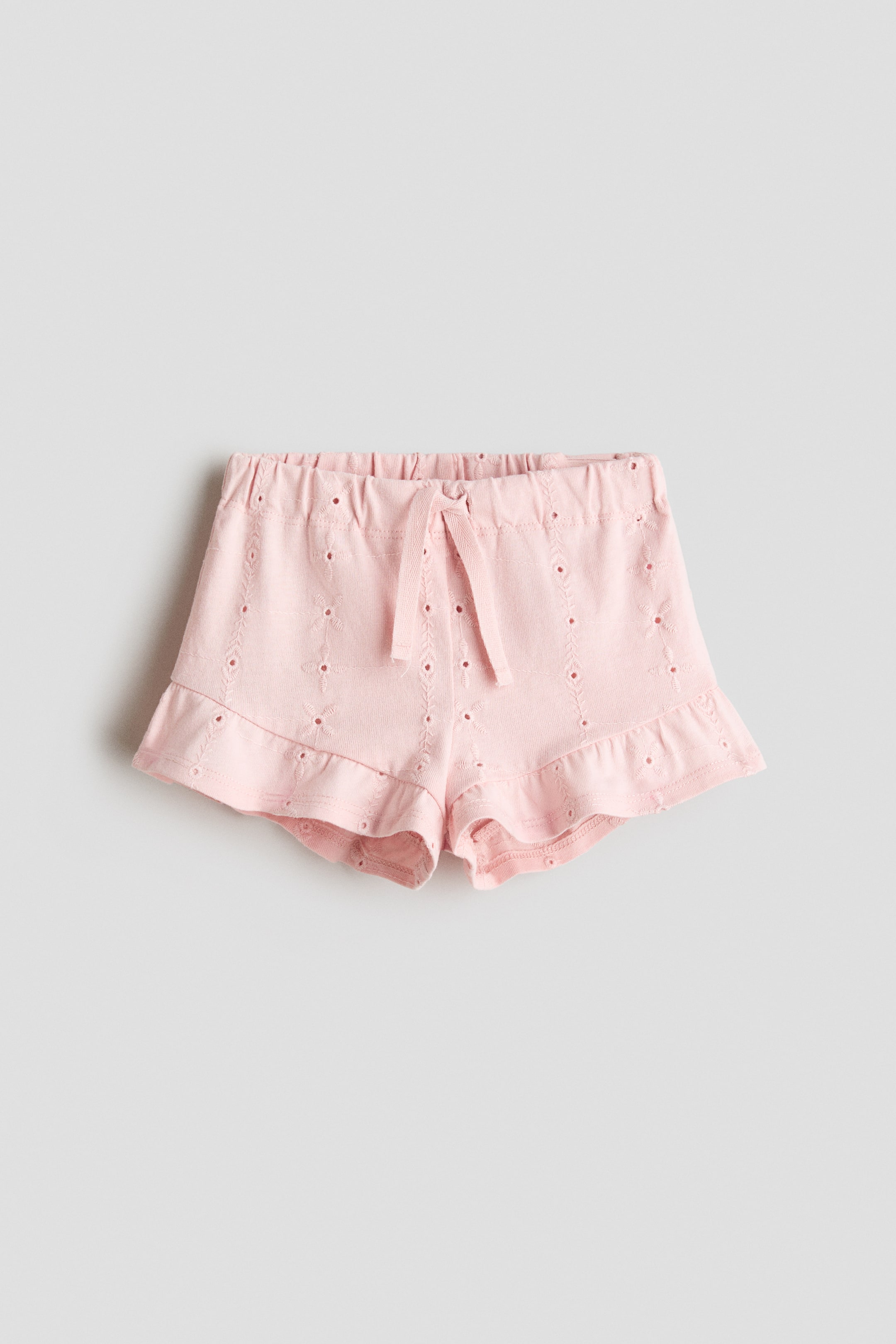 Agrandir l'image: Shorts roses présentés de face, dotés d'une taille élastiquée avec un cordon décoratif noué en nœud, des détails en broderie anglaise sur l'ensemble du tissu jersey de coton doux, et des ourlets ornés de volants.