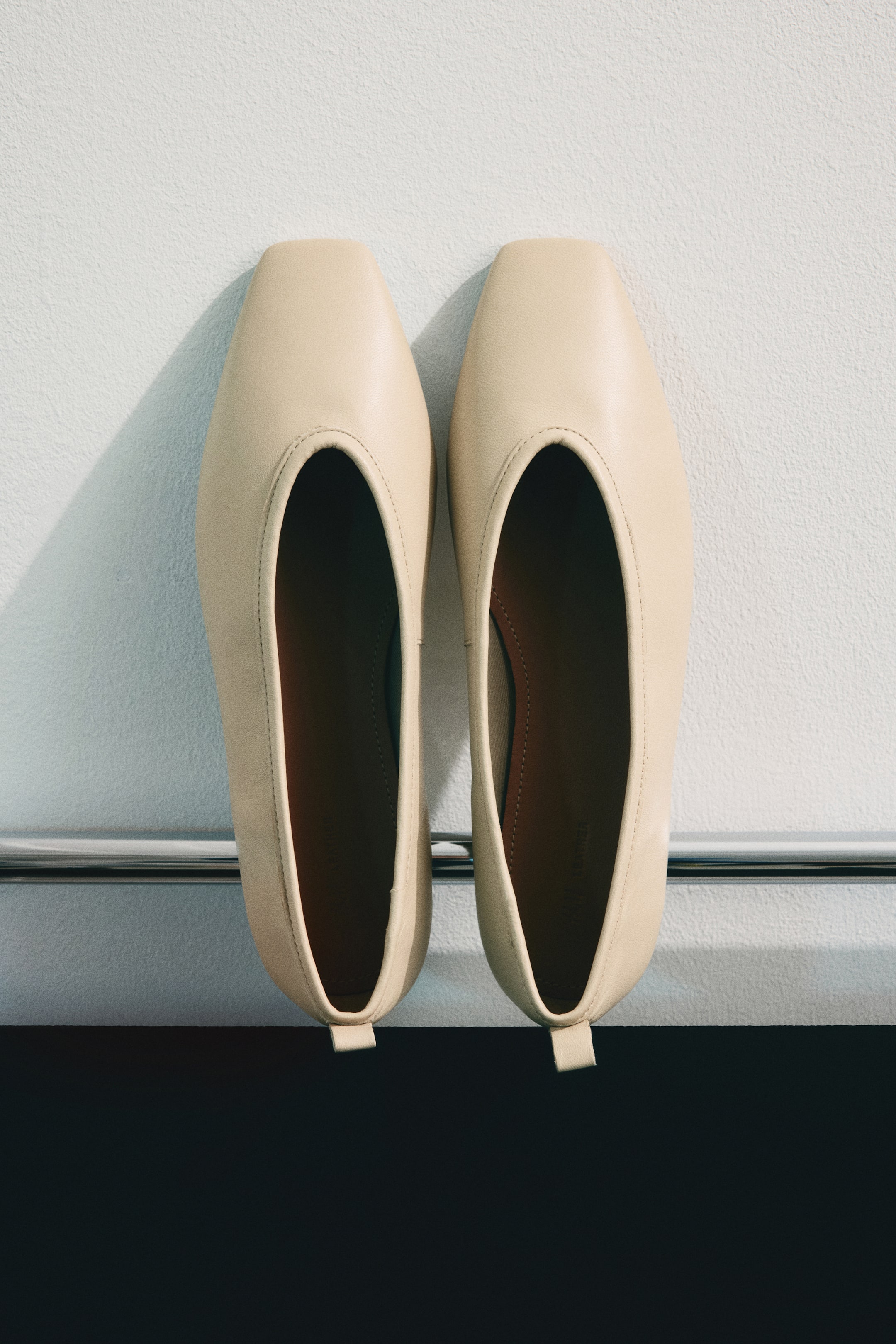 查看大圖: 兩雙淺米色方頭芭蕾舞平底鞋並排擺放，鞋頭朝上。每雙鞋的鞋口處都有明顯的縫線，深棕色鞋墊，以及後跟處的小拉環。