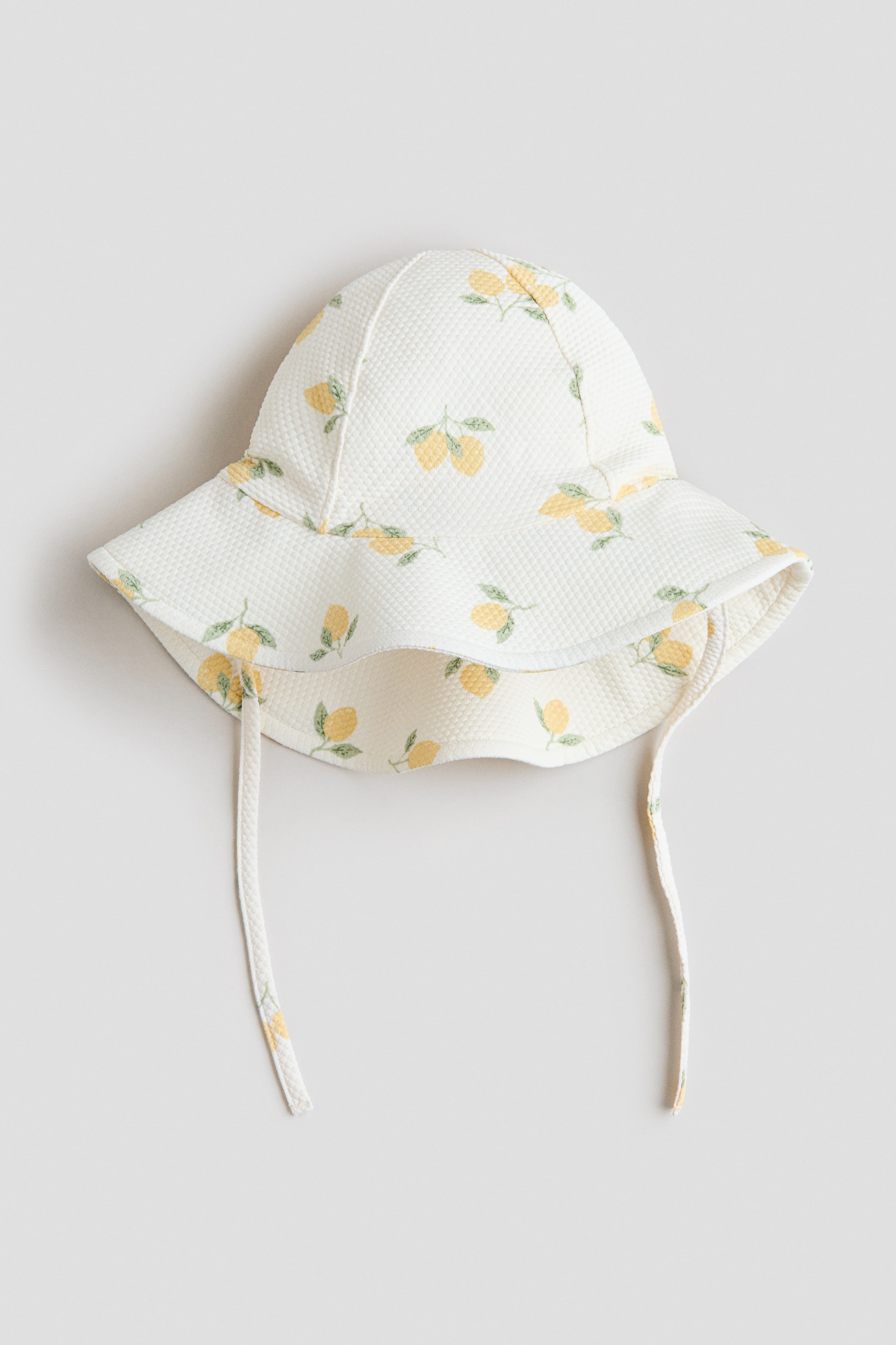 View larger image: A sun hat is showcased from the front, crafted in a cream-coloured textured fabric adorned with a yellow lemon and green leaf motif. It boasts a wide, floppy brim that extends longer at the back, accompanied by matching fabric ties on either side.