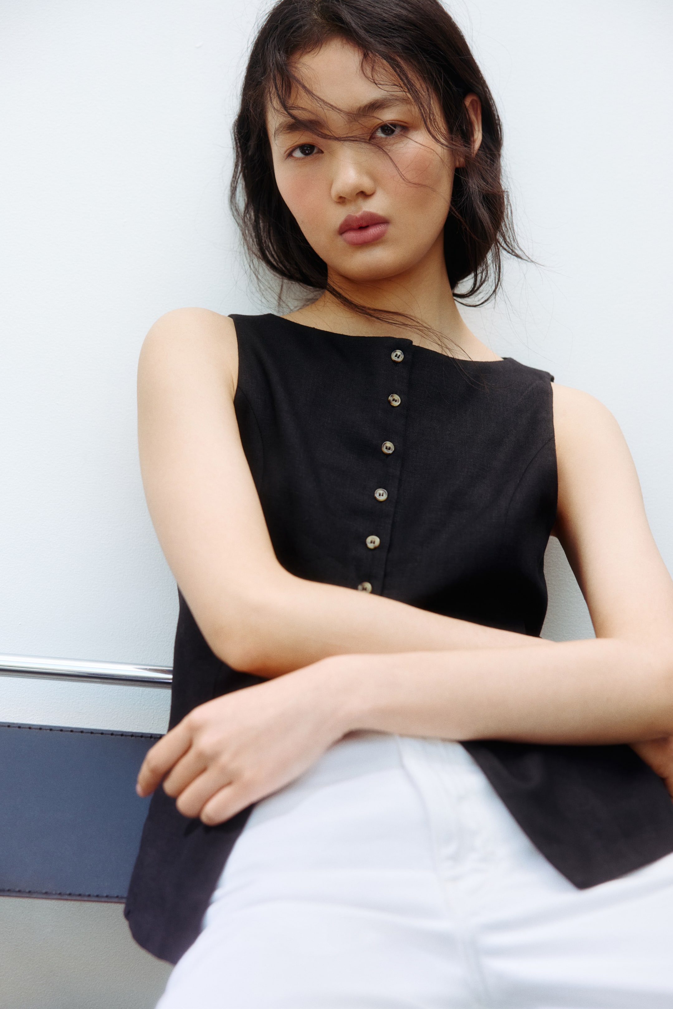 View larger image: A black sleeveless top featuring a straight neckline and a front button placket adorned with multiple light-coloured buttons. The textured fabric offers a relaxed fit, perfectly complementing white trousers.