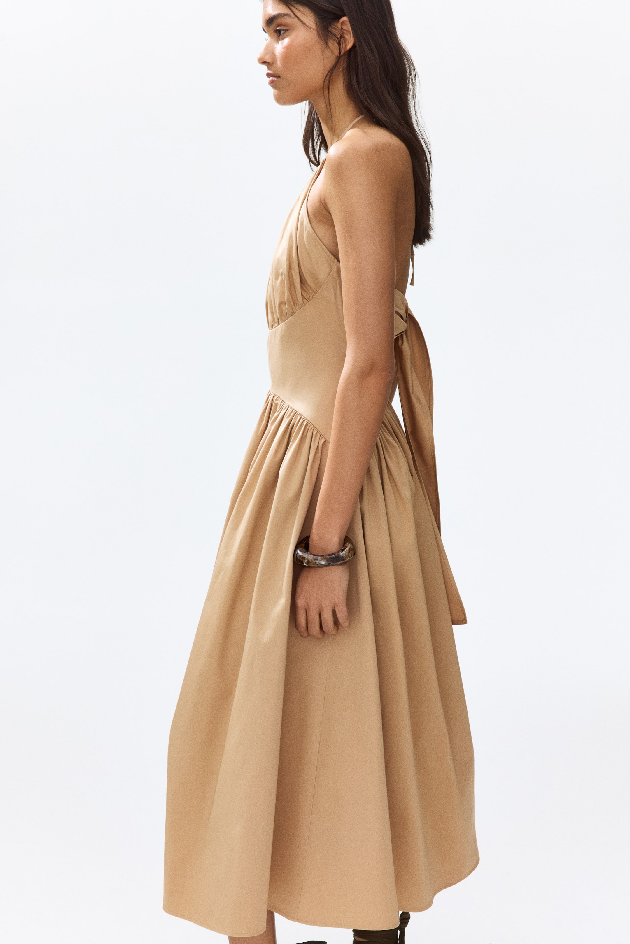 View larger image: The tan halter neck dress showcases a ruched bodice, a fitted waist, and a full, gathered skirt. The halter straps tie at the back of the neck, and a tie detail forms a bow at the back waist, visible from a side profile.