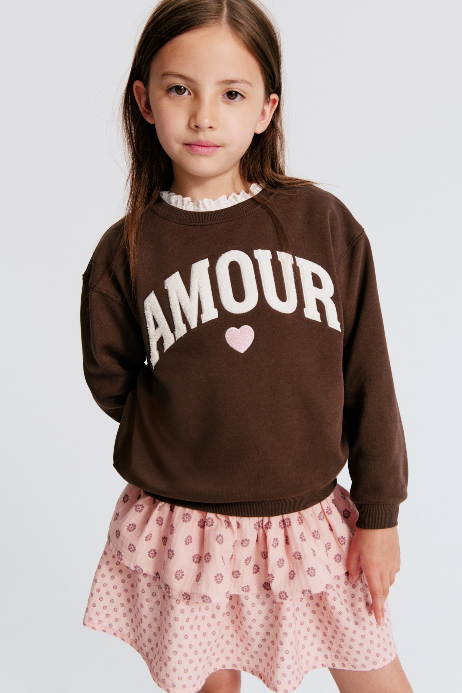 Een bruine sweatshirt met "AMOUR" in witte, gestructureerde letters en een roze hart op de voorkant, met een ronde geribde halslijn, verlaagde schouders en geribde boorden en zoom. - 1