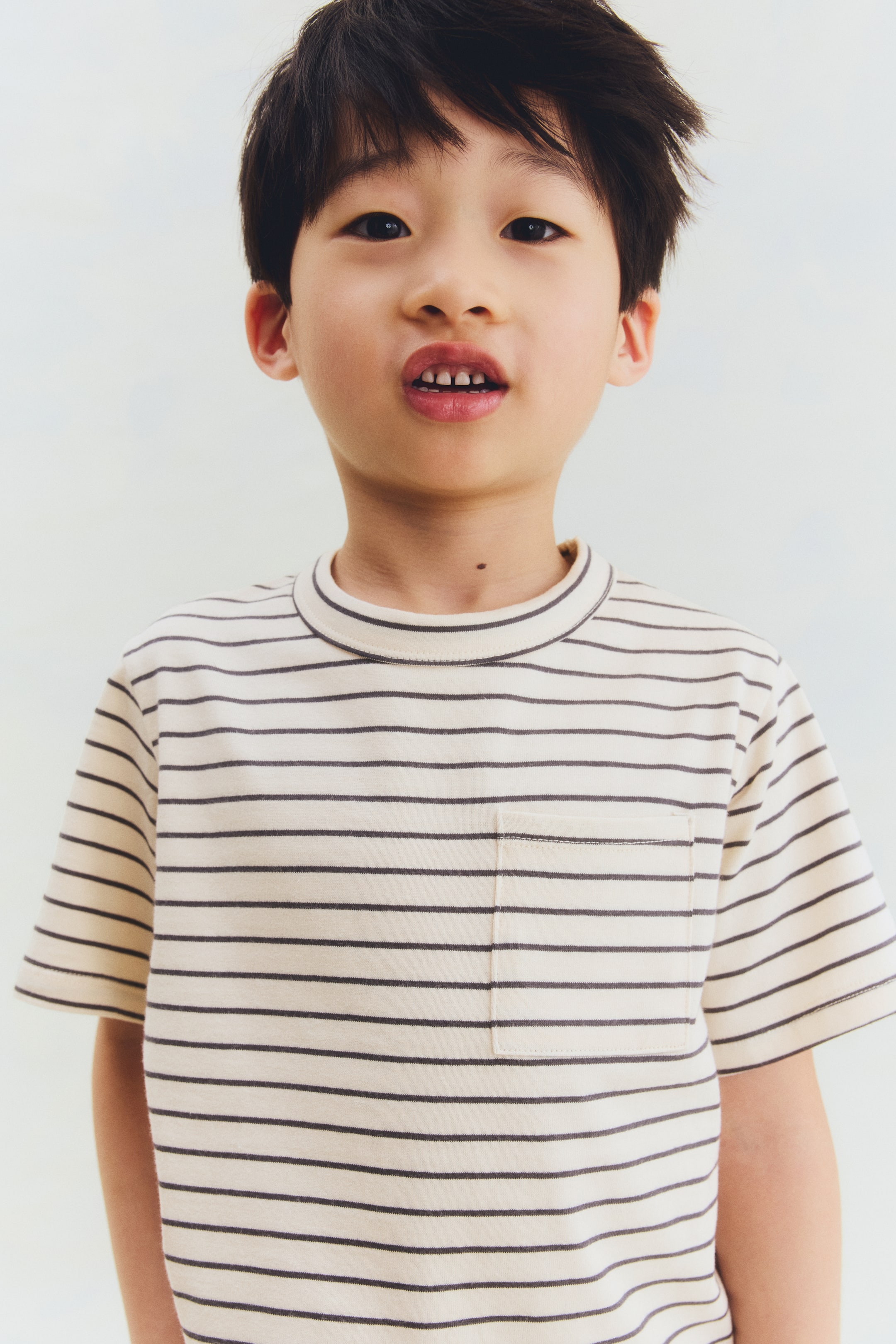 Agrandir l'image: Un t-shirt crème avec des rayures horizontales foncées est présenté de face, mettant en valeur un col rond côtelé et une poche plaquée sur la poitrine gauche.