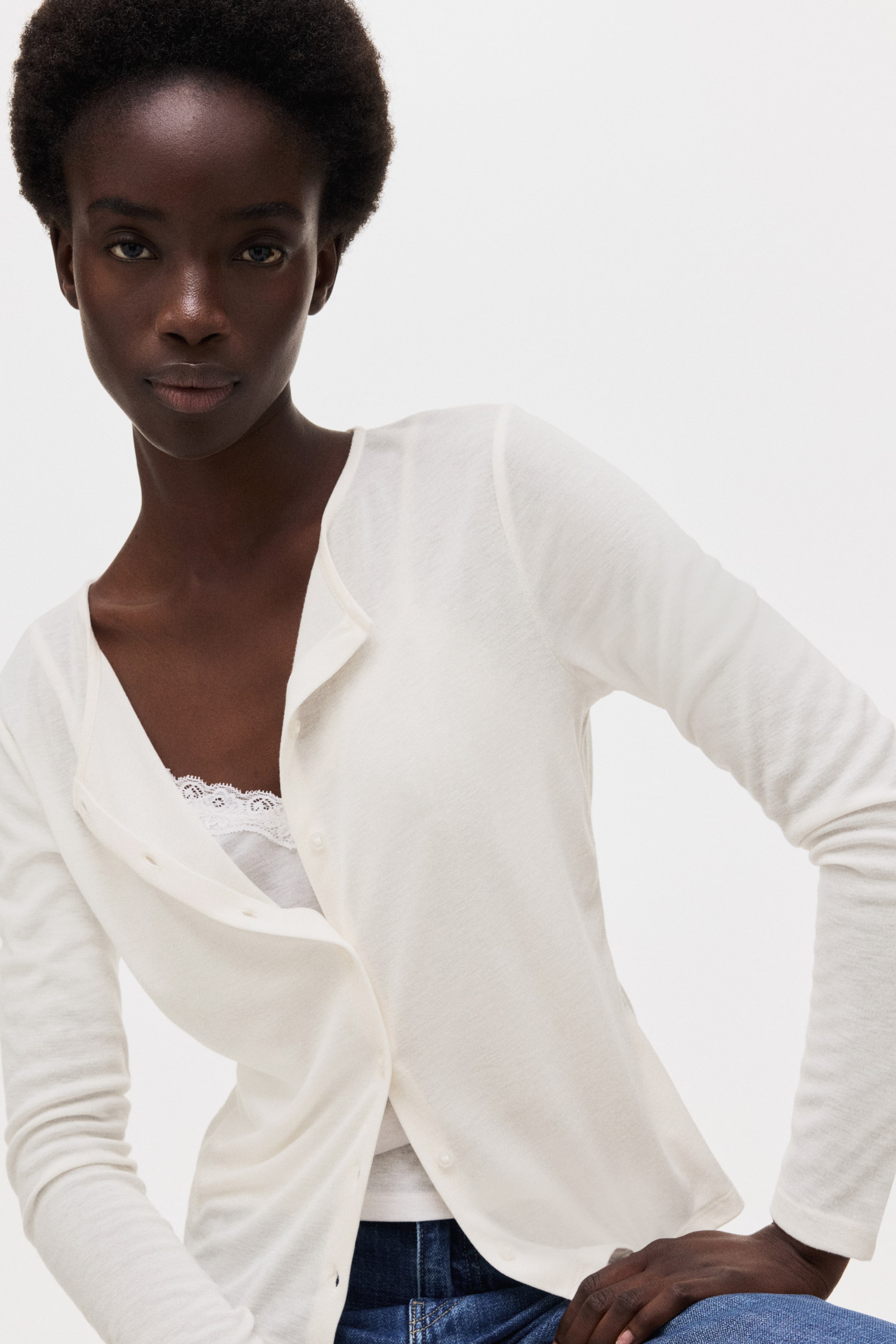 View larger image: A white, lightweight cardigan is being worn, featuring a V-neck and long sleeves. It is partially buttoned down the front with small, pearl-like buttons, revealing a white lace-trimmed camisole underneath. The cardigan's hem curves, and its fitted silhouette is visible as it angles slightly forward.