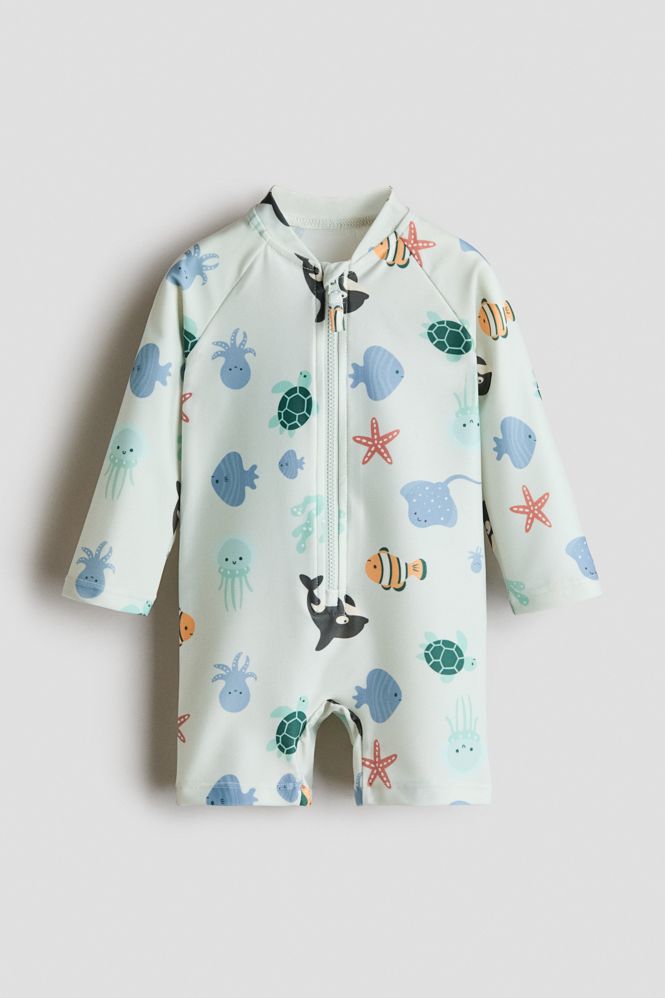 Ampliar la imagen: Un traje de baño estampado se muestra de frente, con una base verde claro y un patrón colorido de criaturas marinas. Está diseñado con un pequeño cuello alto, mangas largas raglán y piernas cortas, y se cierra al frente con un cierre y protector de barbilla.