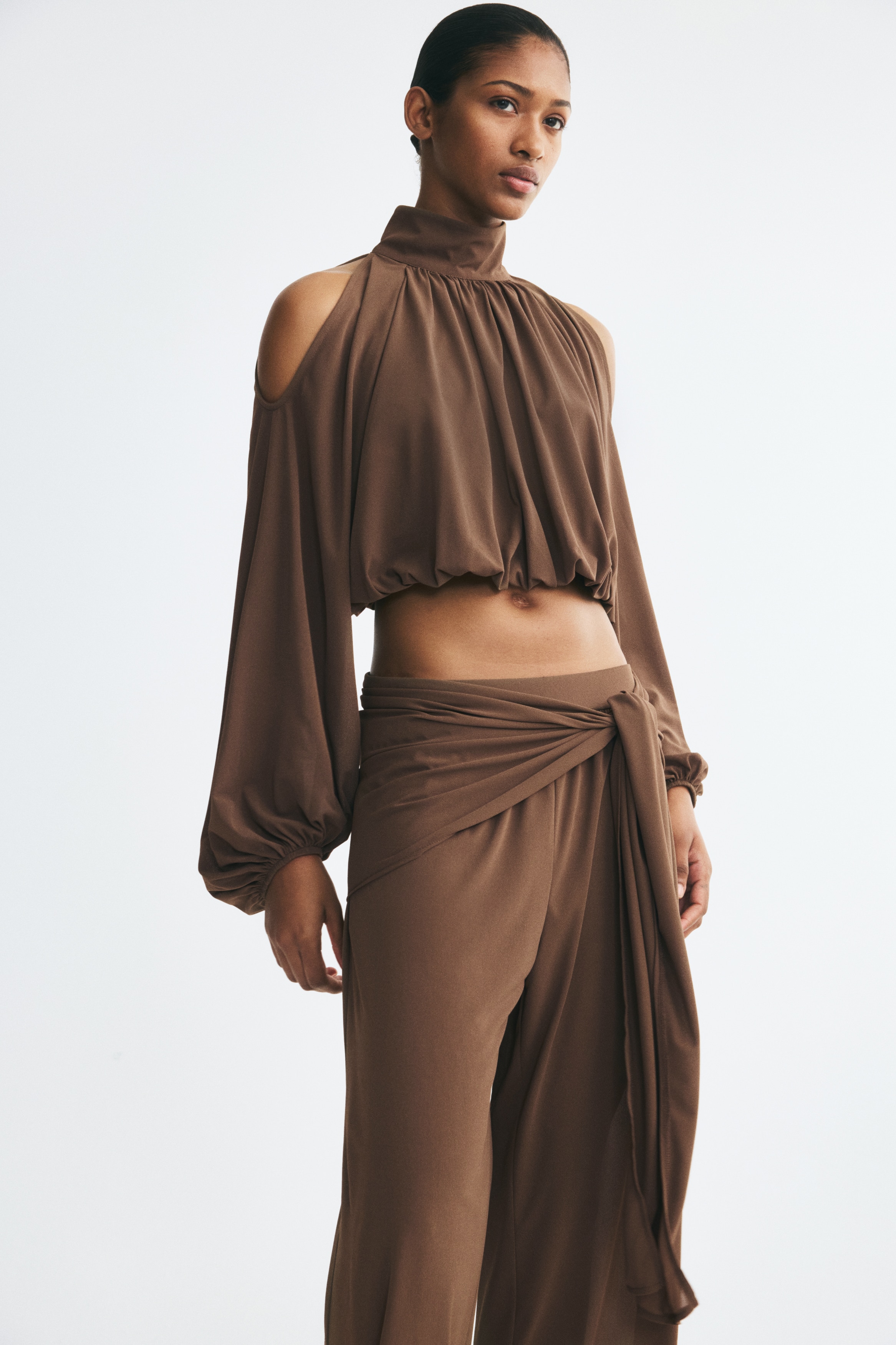 Brown wide-leg pants are shown from the front, featuring a high-rise waist with a draped fabric overlay wrapping around the hips and tied in a loose knot at the center. The fluid, lightweight fabric falls into a relaxed, wide-leg silhouette.