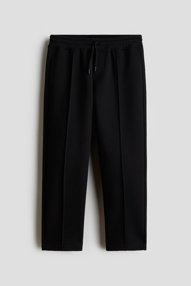 A pair of black straight-leg trousers is shown from the front, featuring an elasticated waistband with an adjustable drawstring. The trousers are crafted from a textured knit fabric and feature prominent pintuck seams running down the centre of each leg to create a structured look. - 1