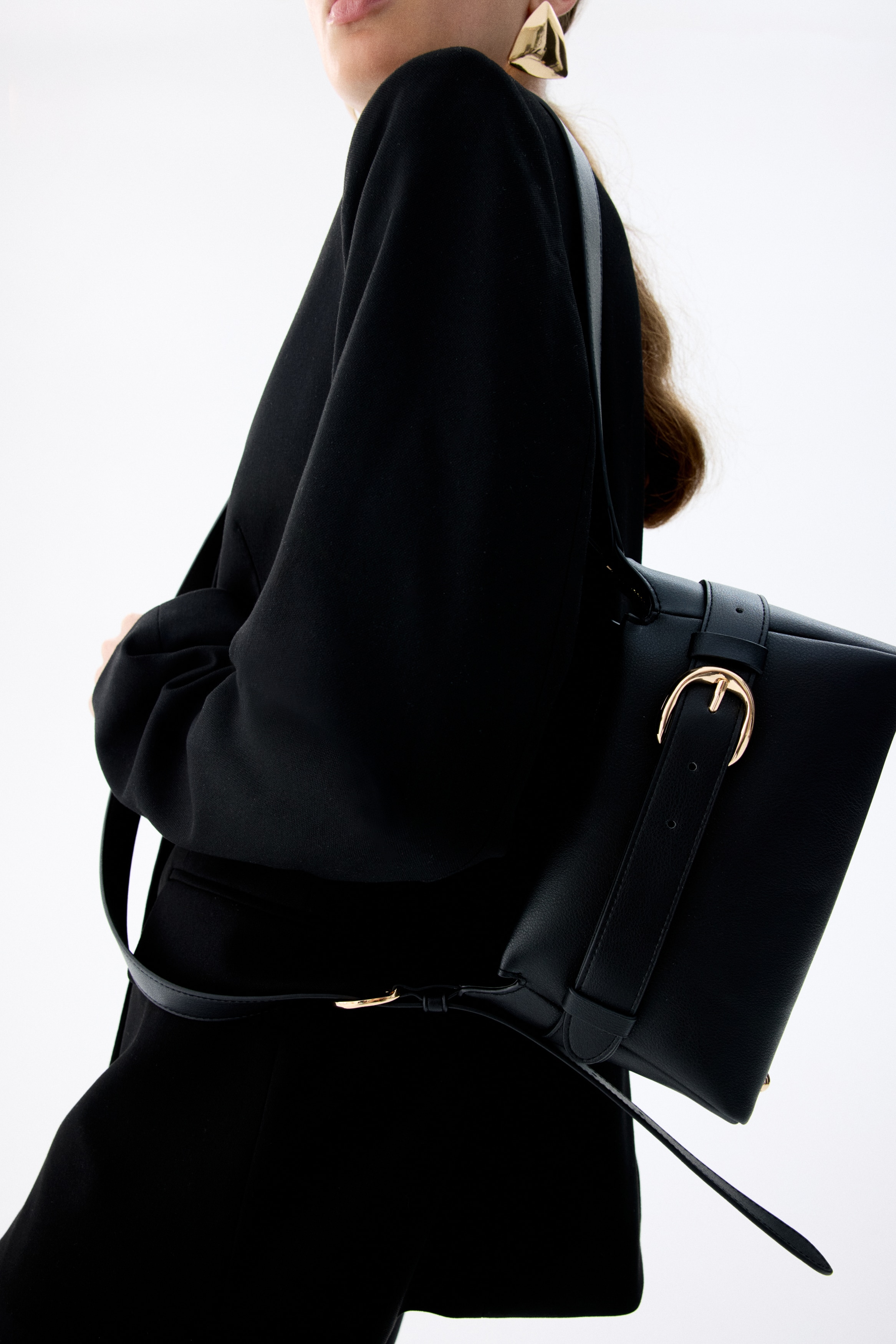 A black crossbody bag is hanging, showcasing its front view. It is made of coated fabric, featuring an adjustable shoulder strap and a zip at the top. A belt-look strap is wrapping around the bag and fastening with a gold-toned metal buckle at the front.