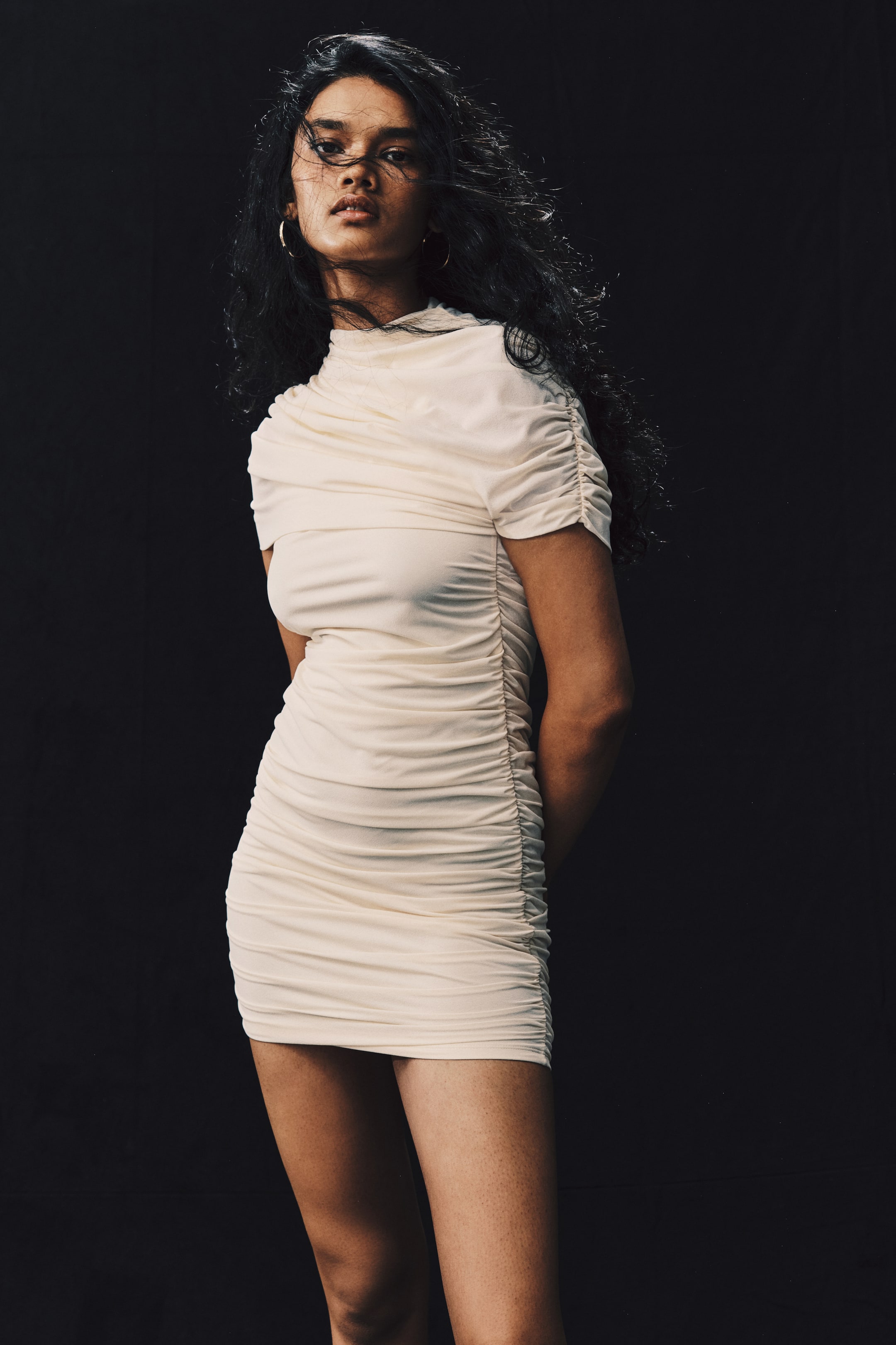 View larger image: The model is wearing a cream-colored fitted mini dress in textured jersey, featuring extensive ruching and draped gathers. The dress has a high stand collar, an asymmetrical draped cape layer over the upper body, and a gathered short sleeve.