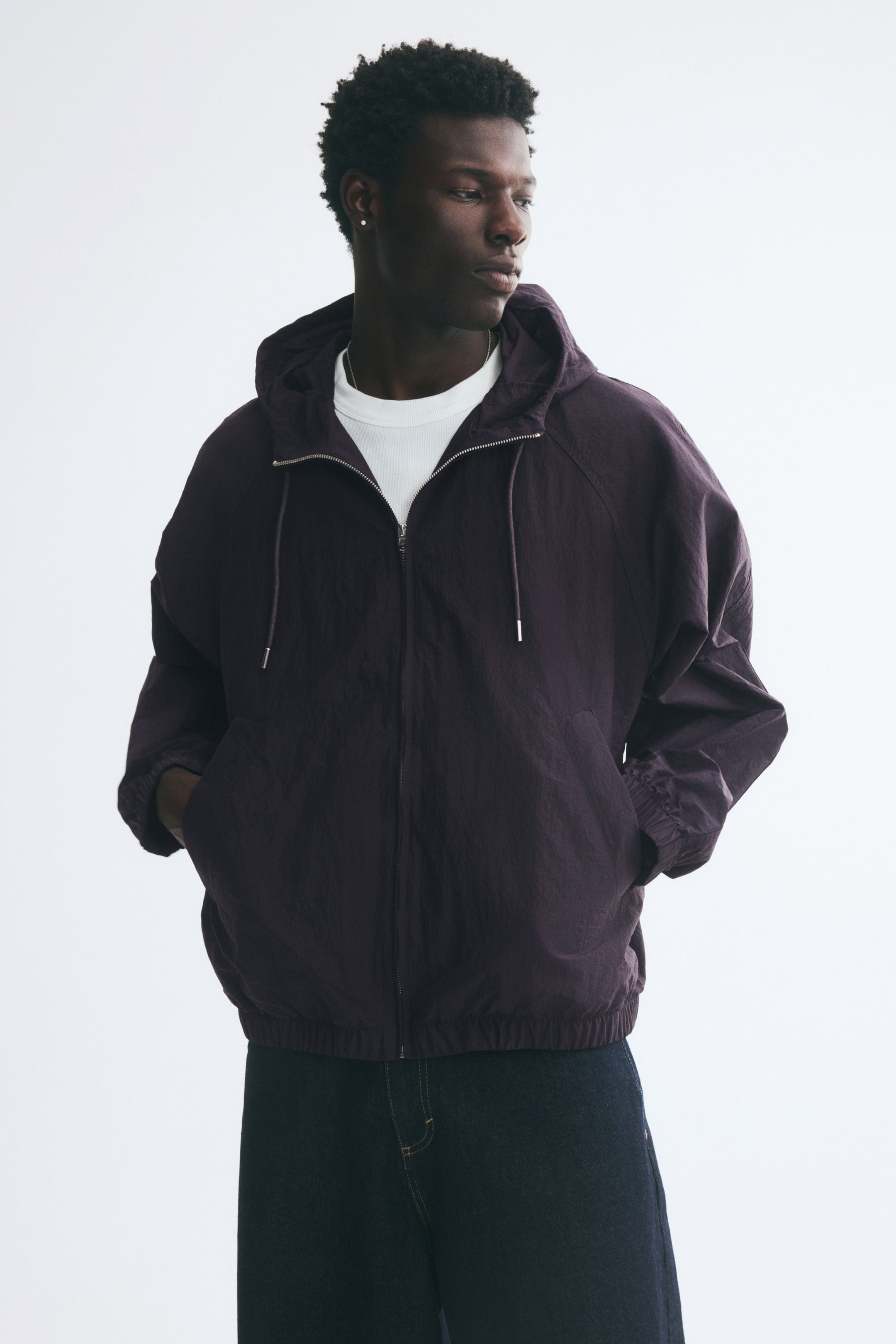 View larger image: A dark purple nylon jacket with a drawstring hood, raglan sleeves, and an elasticated hem is being worn by the model. The jacket is zipped up over a white T-shirt, and the model has their hands placed inside the front welt pockets.