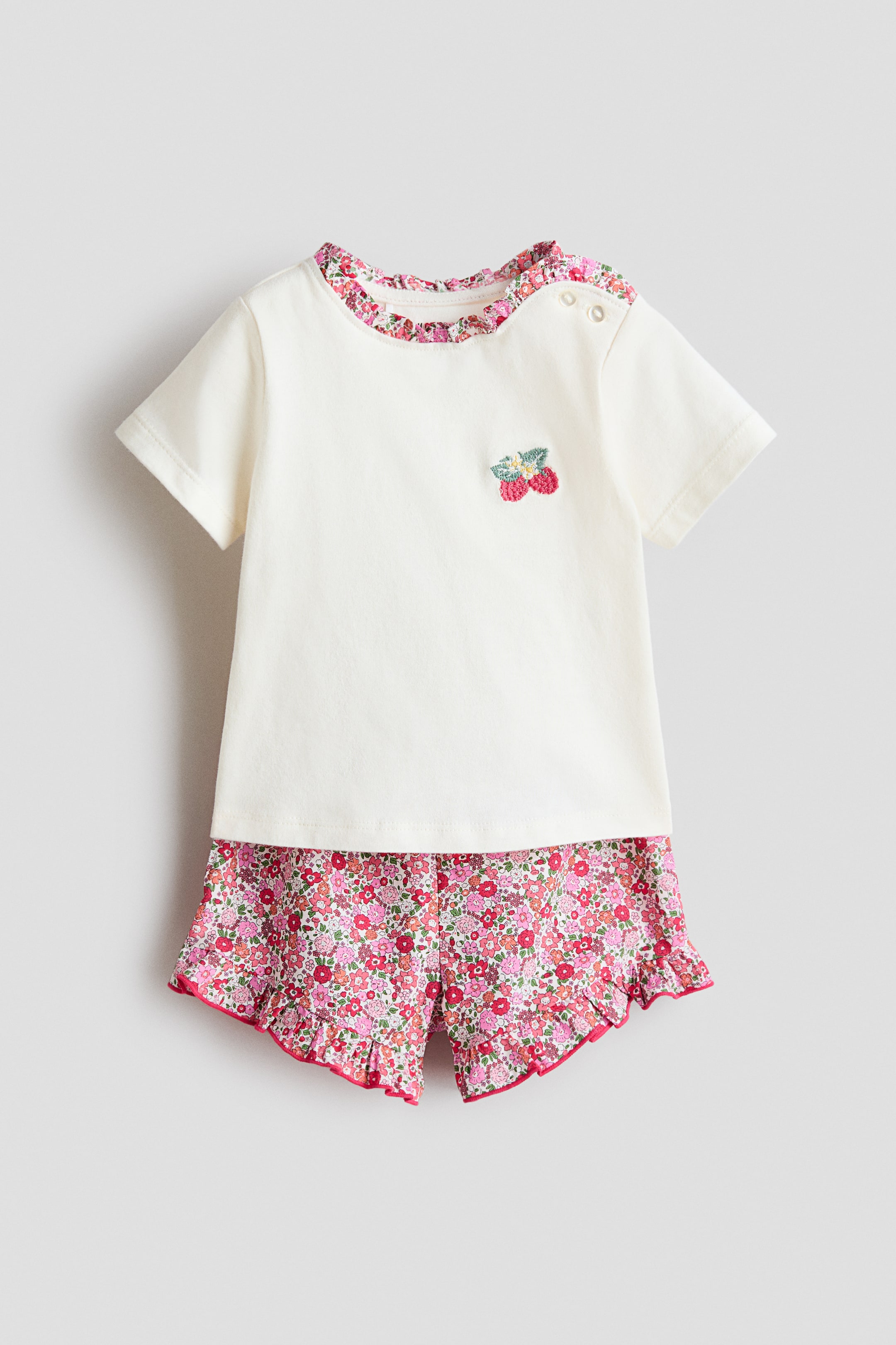View larger image: A two-piece baby outfit is shown from the front. The cream short-sleeved top features a ruffled floral neckline trim, two snap buttons on the left shoulder, and an embroidered berry design on the chest. Matching floral shorts display ruffled hems with pink piping.