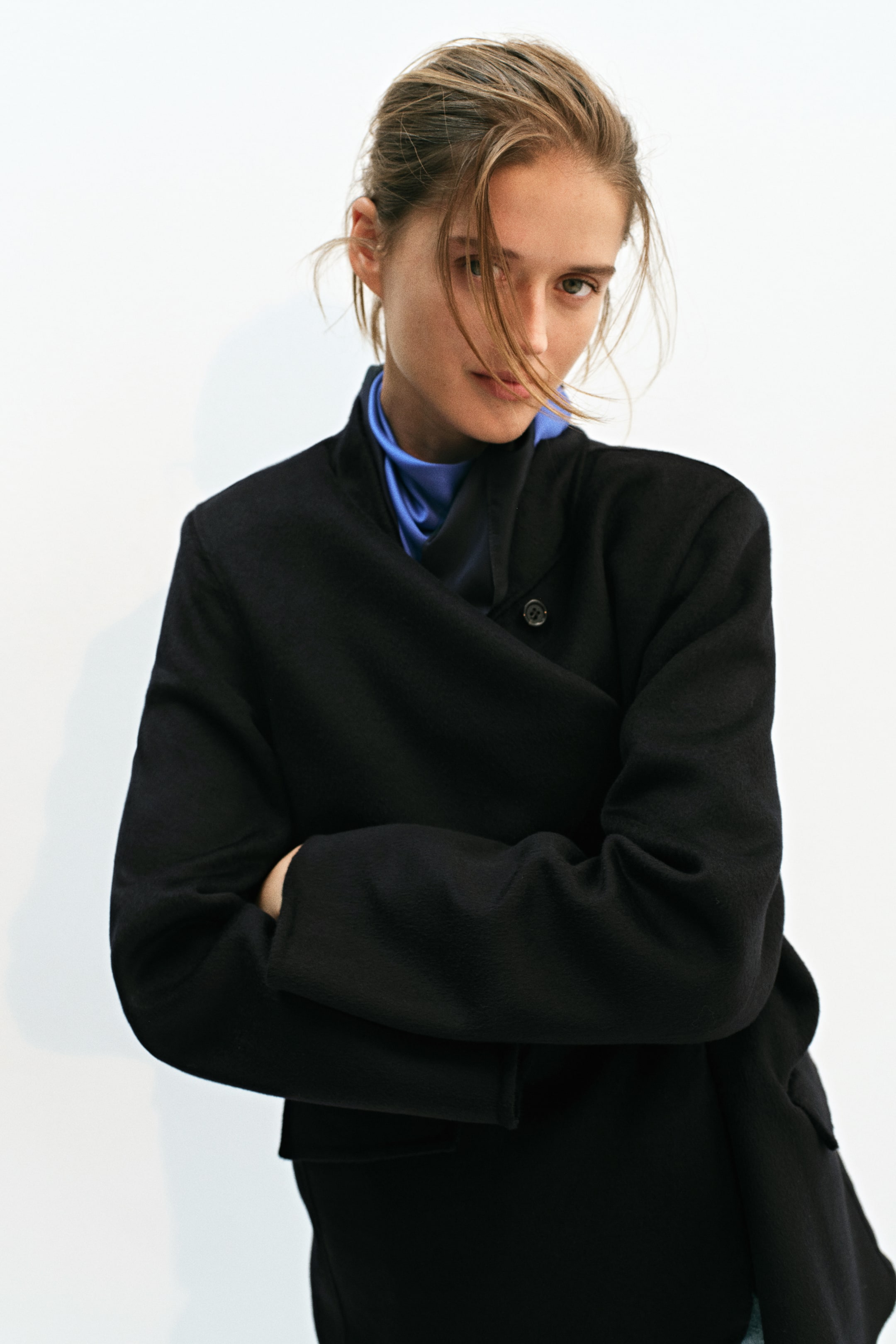 Büyük resmi görüntüle: Keten görünümlü yün karışımından siyah, tek sıra düğmeli bir blazer, kolları çaprazlanmış şekilde model tarafından giyiliyor. Blazer, dik yakaya ve tek görünen düğmeli, hafif kaydırılmış ön açıklığa sahip olup mavi ve siyah saten bir fuların üzerine katmanlanmıştır.