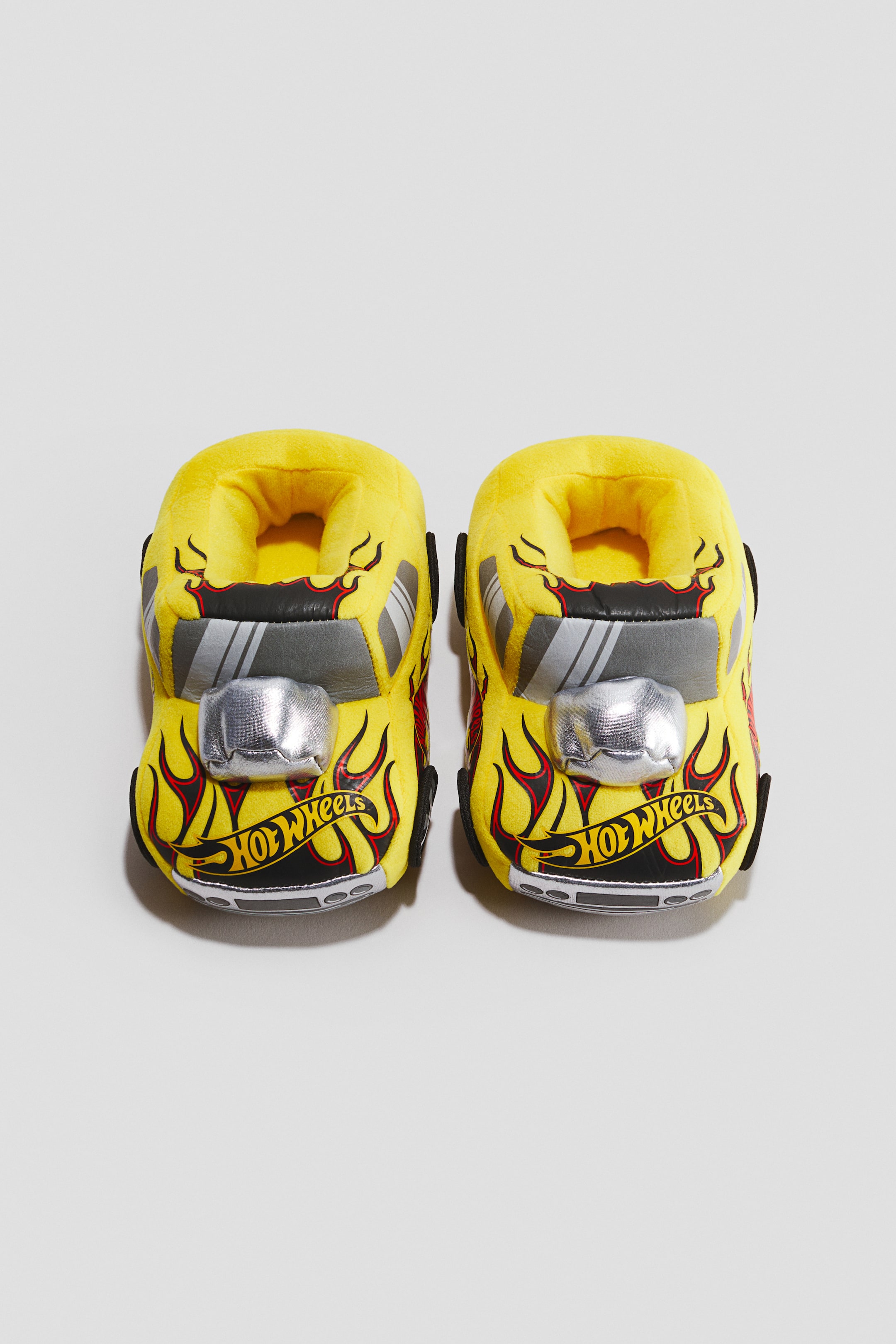 View larger image: A pair of padded, car-shaped slippers in yellow plush fabric is facing front, showcasing a Hot Wheels race car design. The slippers feature black and red flame graphics, a metallic silver plush engine block on the hood, and the "Hot Wheels" logo printed on the front bumper.