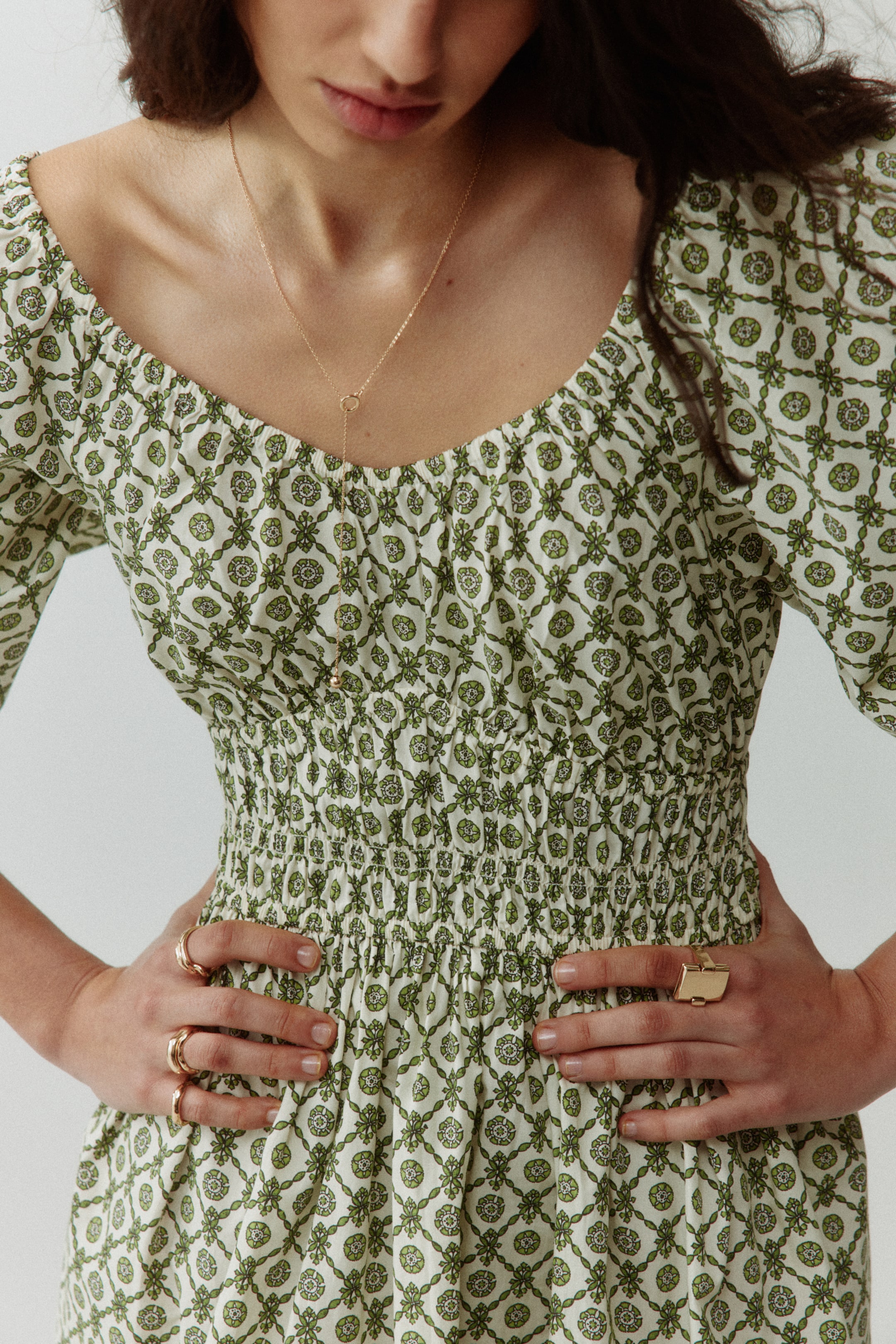 Größeres Bild ansehen: Ein cremefarbenes Kleid mit einem sich wiederholenden grün-blumigen geometrischen Muster verfügt über einen breiten, gerafften Rundhalsausschnitt und umfangreiche Rüschen, die das Oberteil vom Unterbrustbereich bis zur natürlichen Taille verzieren. Eine goldene Y-Halskette ruht auf dem Kleid.