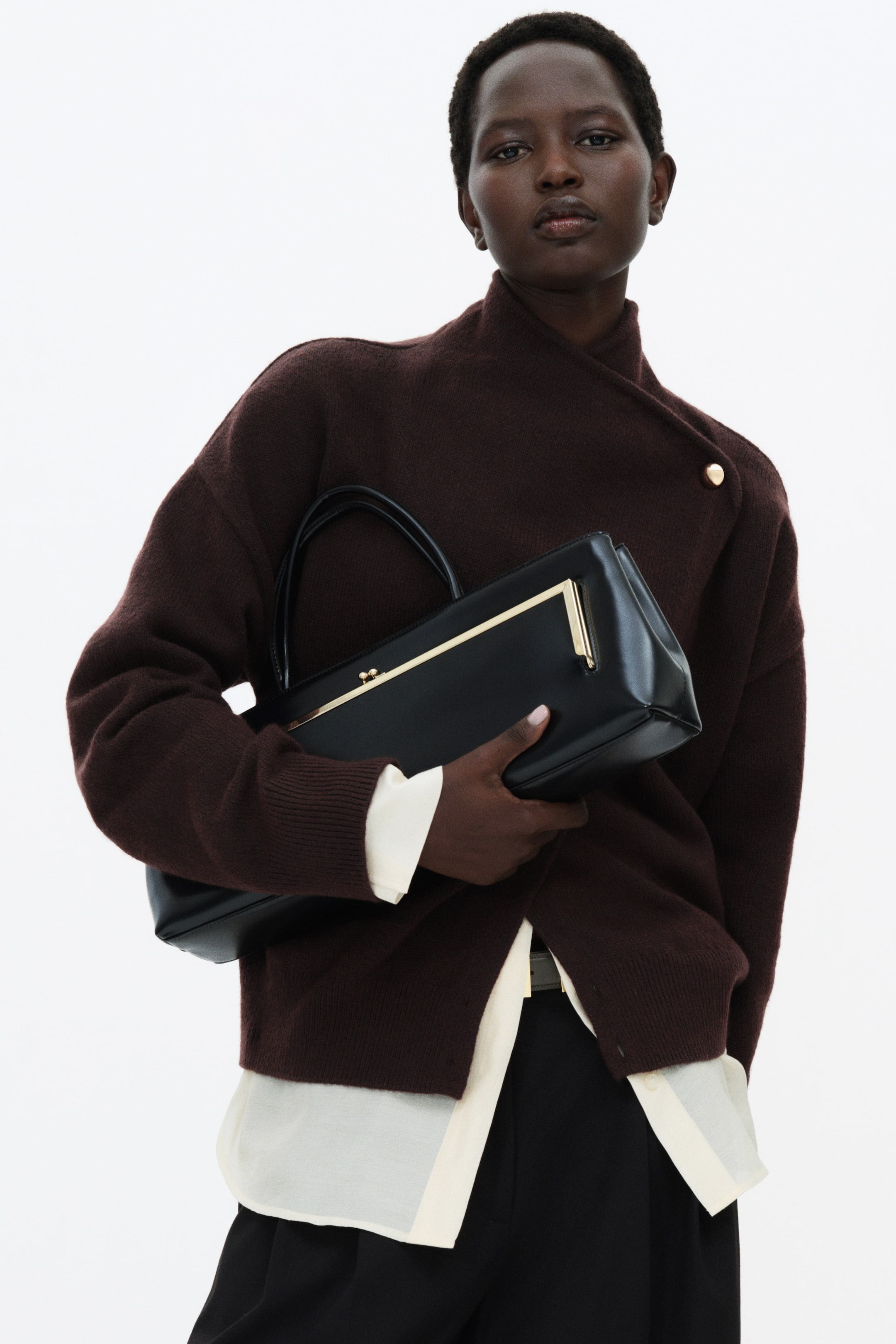 View larger image: A dark brown knit wrap cardigan featuring a high neck and a diagonal wrap front with a single visible gold button is worn by the model. The cardigan is layered over a cream-coloured shirt, and the model is holding a black structured handbag across the front.