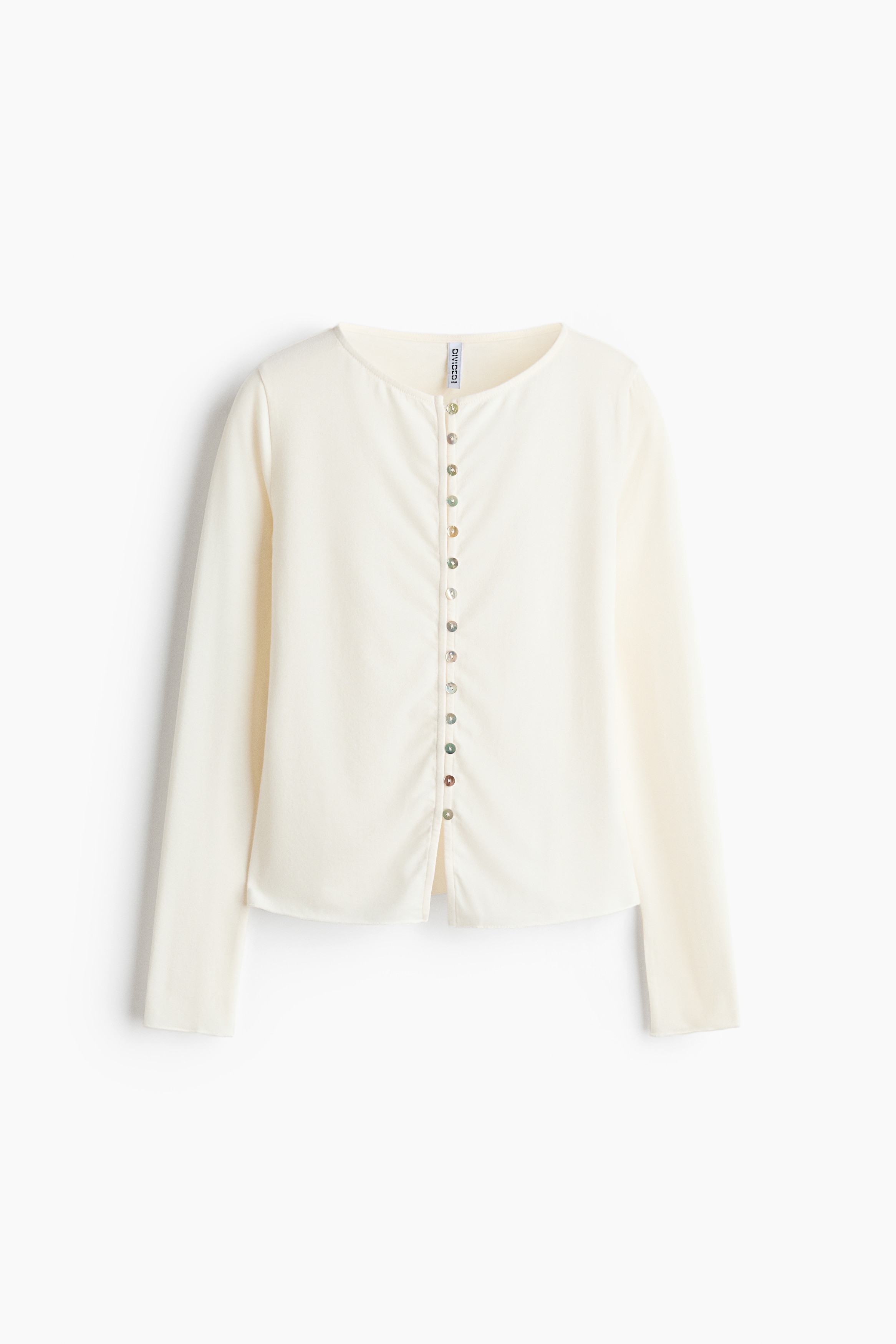 Un top aderente color crema in maglia fine indossato dalla modella, con scollo rotondo, maniche lunghe e chiusura frontale con bottoni e asole. Il top è lasciato parzialmente sbottonato sul bordo inferiore per mettere in risalto lâaddome ed è abbinato a jeans beige a gamba larga.