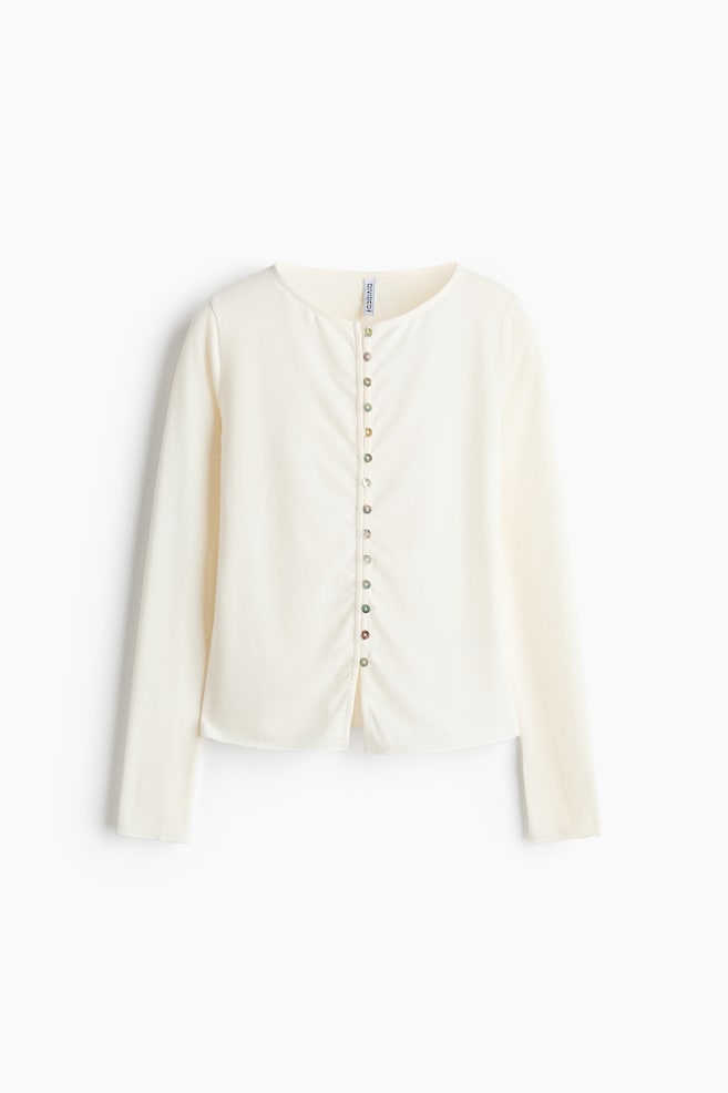 Un top aderente in maglia fine color crema Ã¨ mostrato frontalmente, con scollo rotondo, maniche lunghe e orlo dritto. Una fila verticale di piccoli bottoni in madreperla iridescente chiude il centro davanti con delicati alamari e asole. - 1