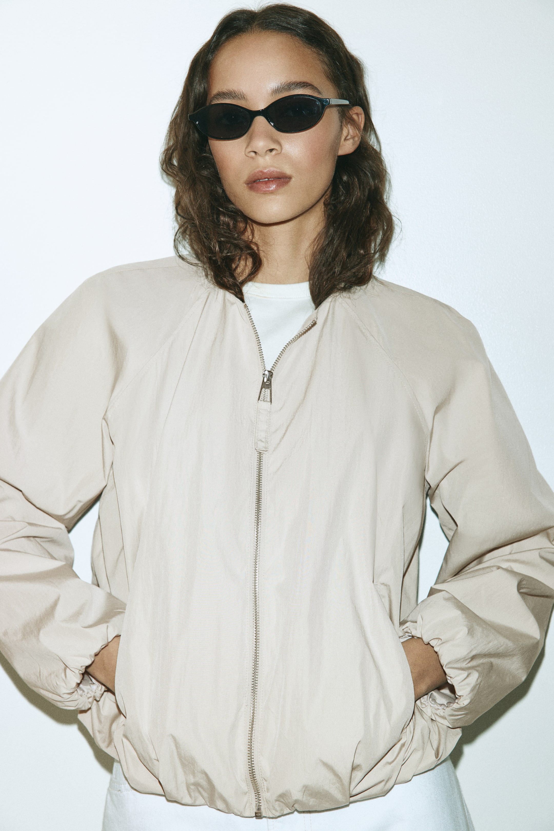 View larger image: A light beige bomber jacket is shown from the front, featuring a full-zip closure with a silver-toned zipper, raglan sleeves, elasticated cuffs and an elasticated hem. It shows side pockets and is worn open over a white T-shirt.