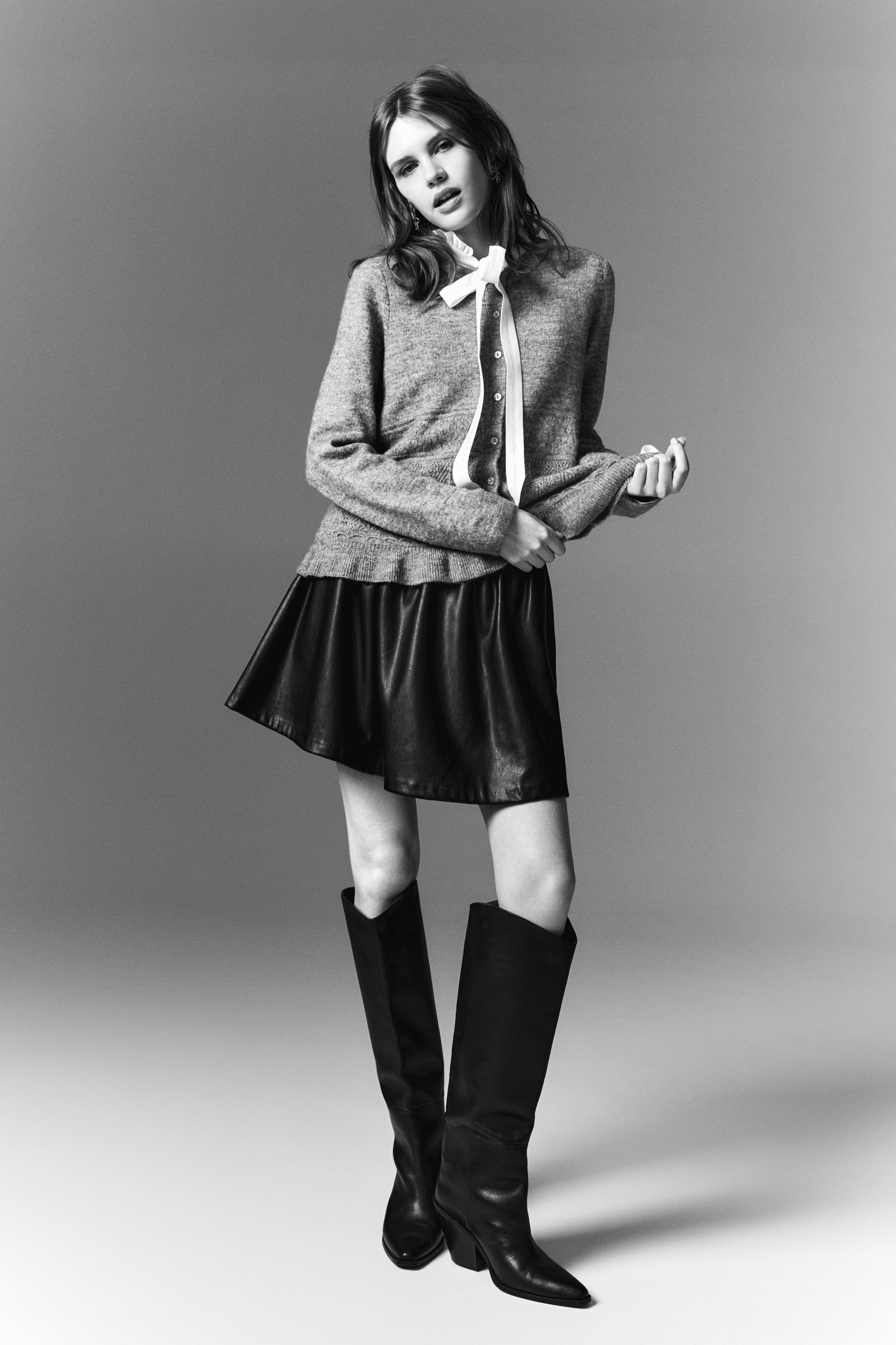 View larger image: A cardigan with a round neckline, buttons down the front, and a frill trim at the hem is being worn by the model. A ruffled blouse with a white ribbon tie is visible underneath, and the model is holding her hands together, adjusting a sleeve.