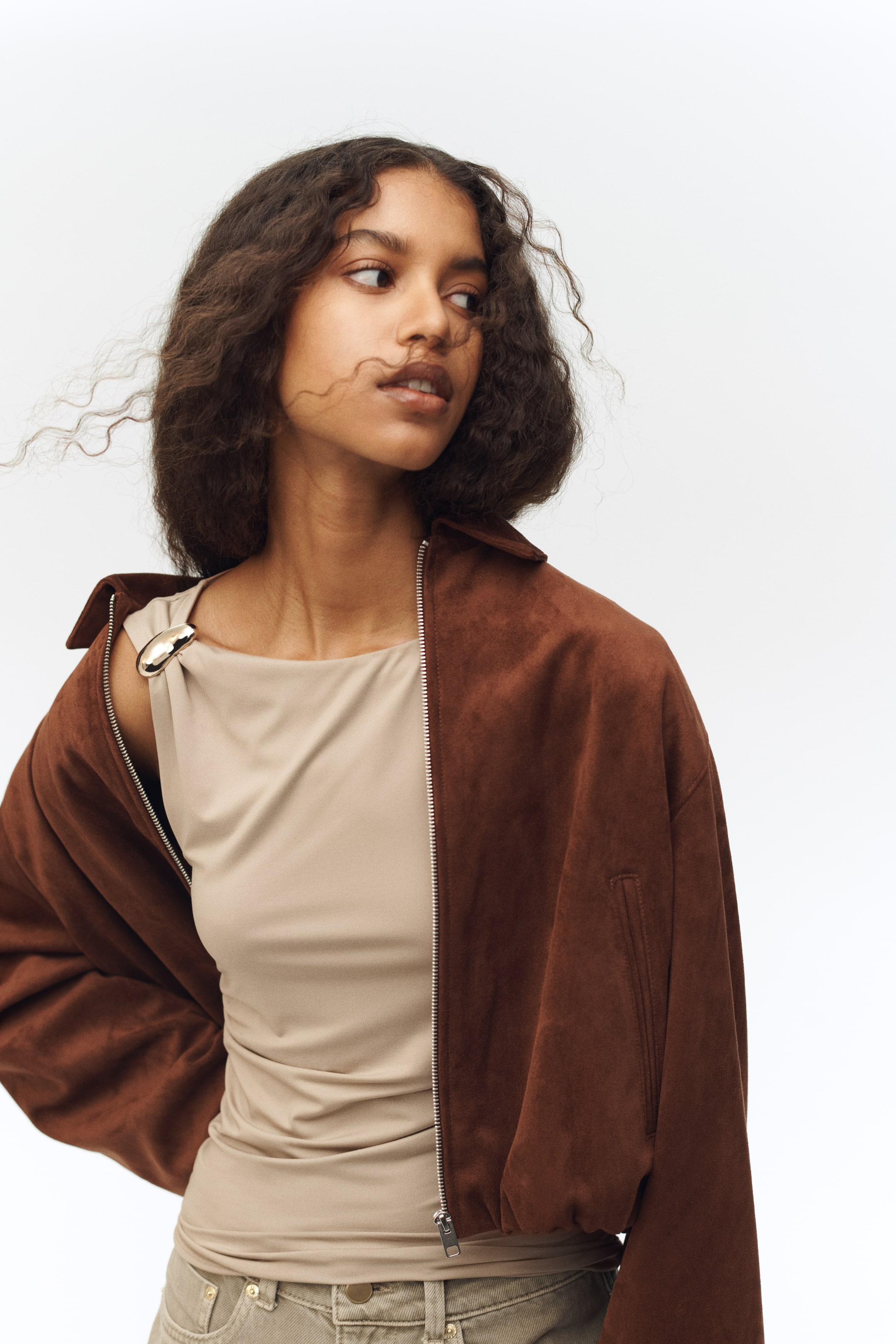 View larger image: A brown suede-like bomber jacket is worn open over a beige draped top, with the jacket's silver-toned zipper visible down the centre. The jacket features a classic collar, dropped shoulders, an angled pocket on the left, and gathered detailing along its hem, while the top showcases an asymmetrical neckline with a metallic oval buckle securing the drape on the left shoulder.