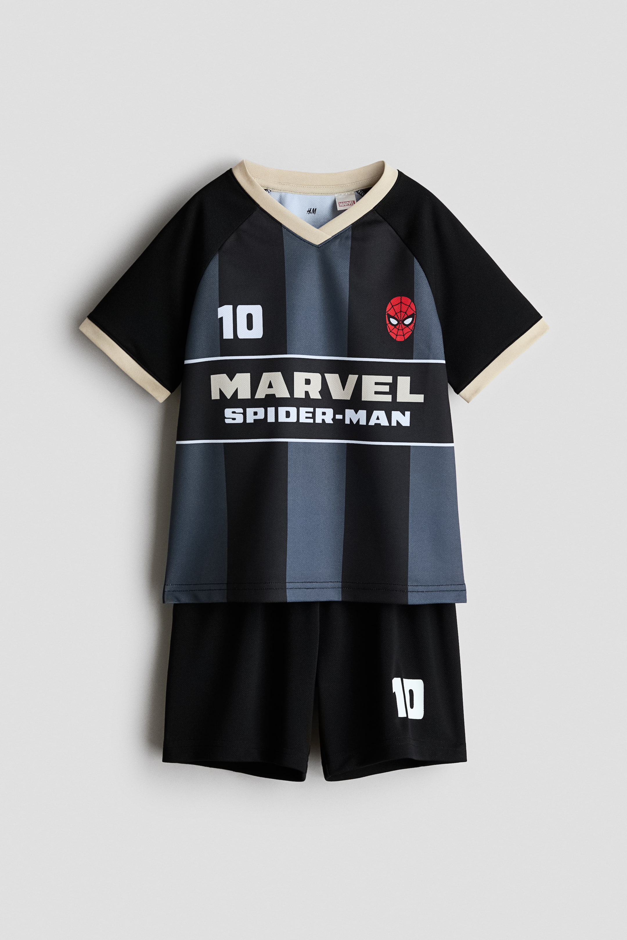 Ingrandisci l'immagine: Di fronte, è esposto un completo da calcio composto da maglia e pantaloncini. La maglia presenta strisce verticali nere e grigie, uno scollo a V beige e rifiniture abbinate sulle maniche corte raglan, con un '10' bianco sul petto sinistro, una maschera di Spiderman rossa sulla destra e il testo 'MARVEL SPIDER-MAN' sul davanti. I pantaloncini neri mostrano una stampa '10' bianca sulla gamba sinistra.