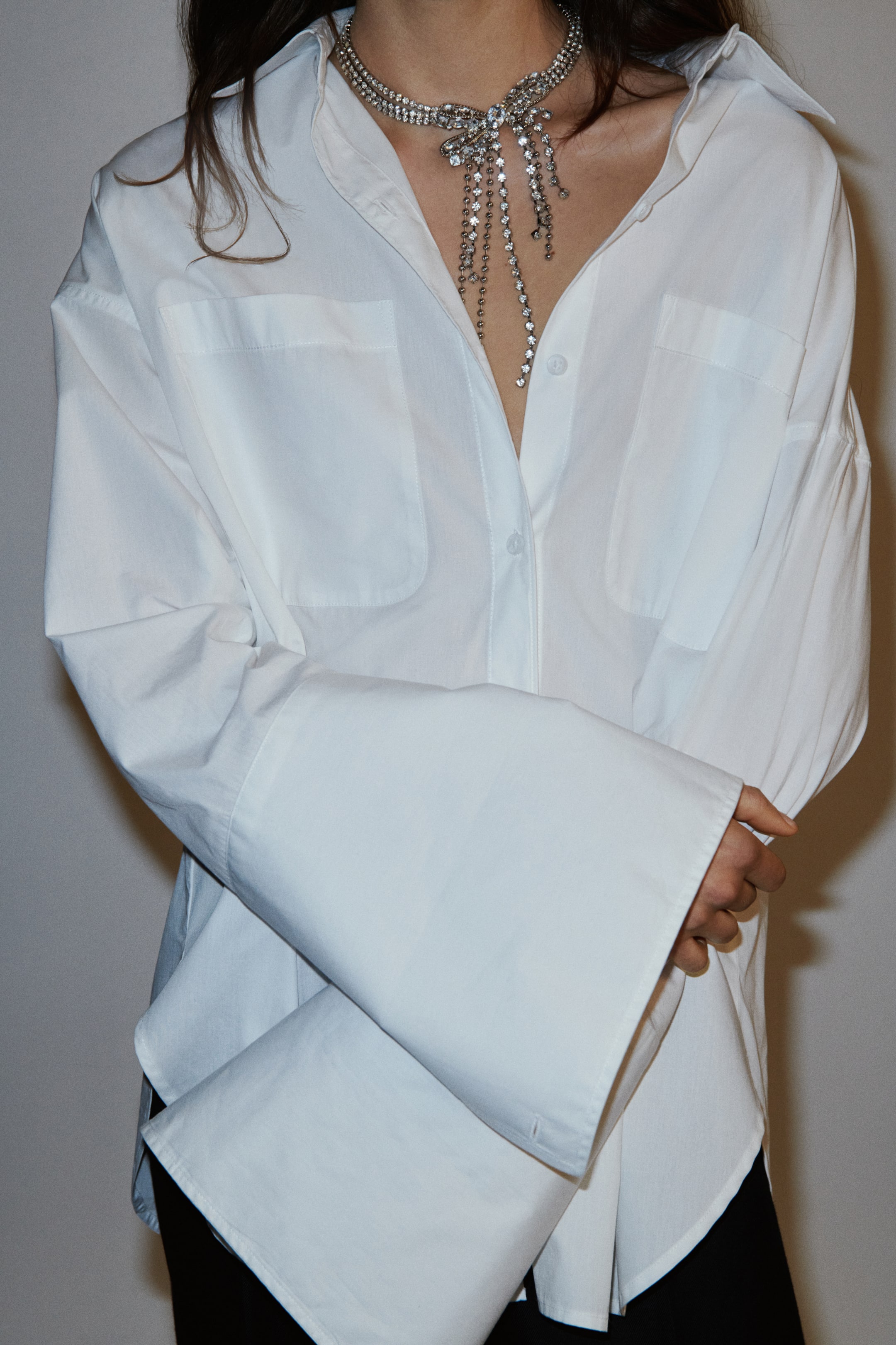 View larger image: A white oversized button-down shirt is being presented from the front, showcasing a classic collar and two front patch pockets. Its wide, long sleeves are extending into broad cuffs, with one cuff appearing folded, and a rhinestone-studded necklace is draping over the shirt's open neckline.