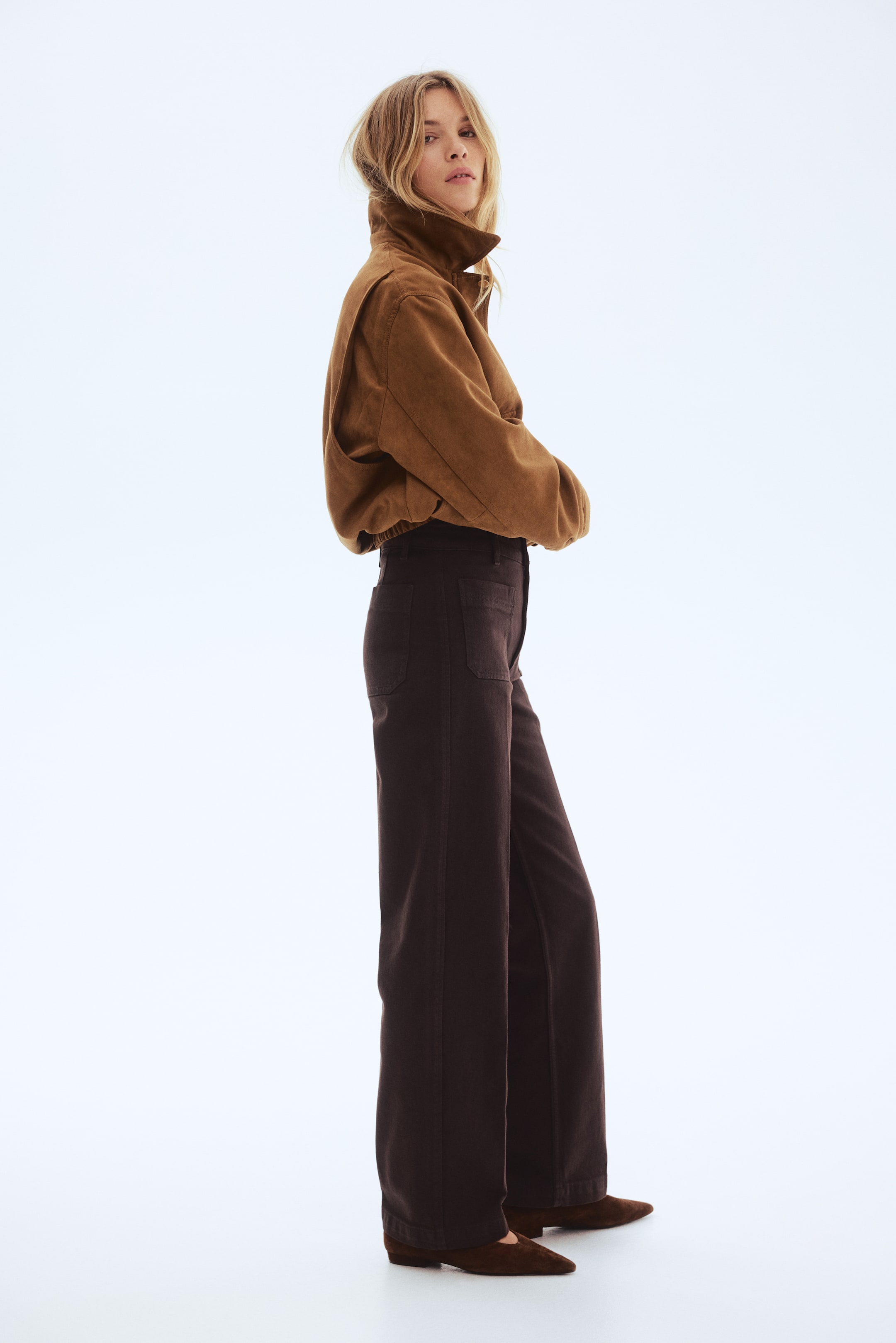 Προβολή μεγαλύτερης εικόνας: Η μοντέλο φοράει τζιν σε σκούρο καφέ χρώμα με φαρδύ πόδι. Τα τζιν διαθέτουν ψηλή μέση με κλείσιμο με κουμπί, τσέπες στο μπροστινό και πίσω μέρος, και πιέτες κατά μήκος του μπροστινού μέρους, με το στρίφωμα να φτάνει στην κορυφή του ποδιού.