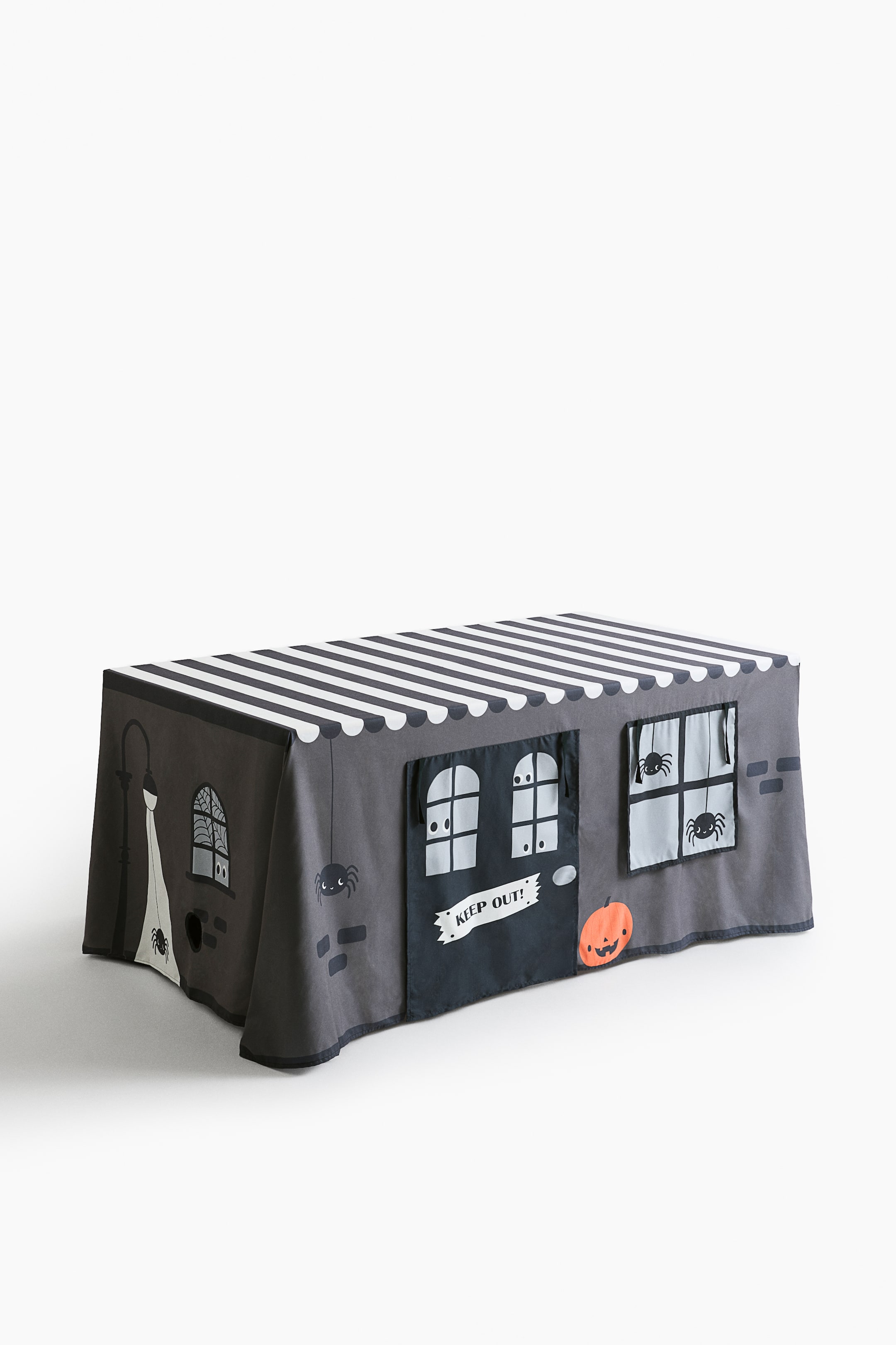 View larger image: A gray woven table tent is shown from a front-left angle, featuring a haunted house motif. The top has a black-and-white striped awning design. The front panel displays a black door with a “KEEP OUT!” sign and a window with spiders, both styled as roll-down curtains. A pumpkin graphic is also visible on the front, while the left side panel shows a lamppost and a spiderweb window.