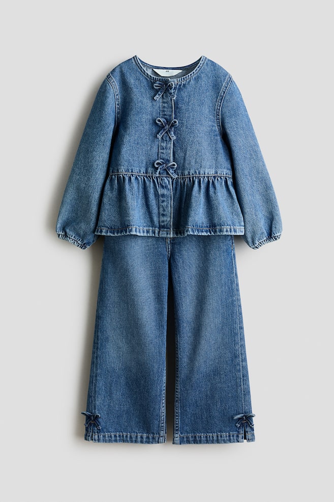 A denim two-piece outfit is shown front-on, featuring a top with a round neck, long sleeves with gathered cuffs and a peplum hem, with three small bows adorning the front placket. The matching wide-leg trousers feature small bows at the outer ankles. - 1