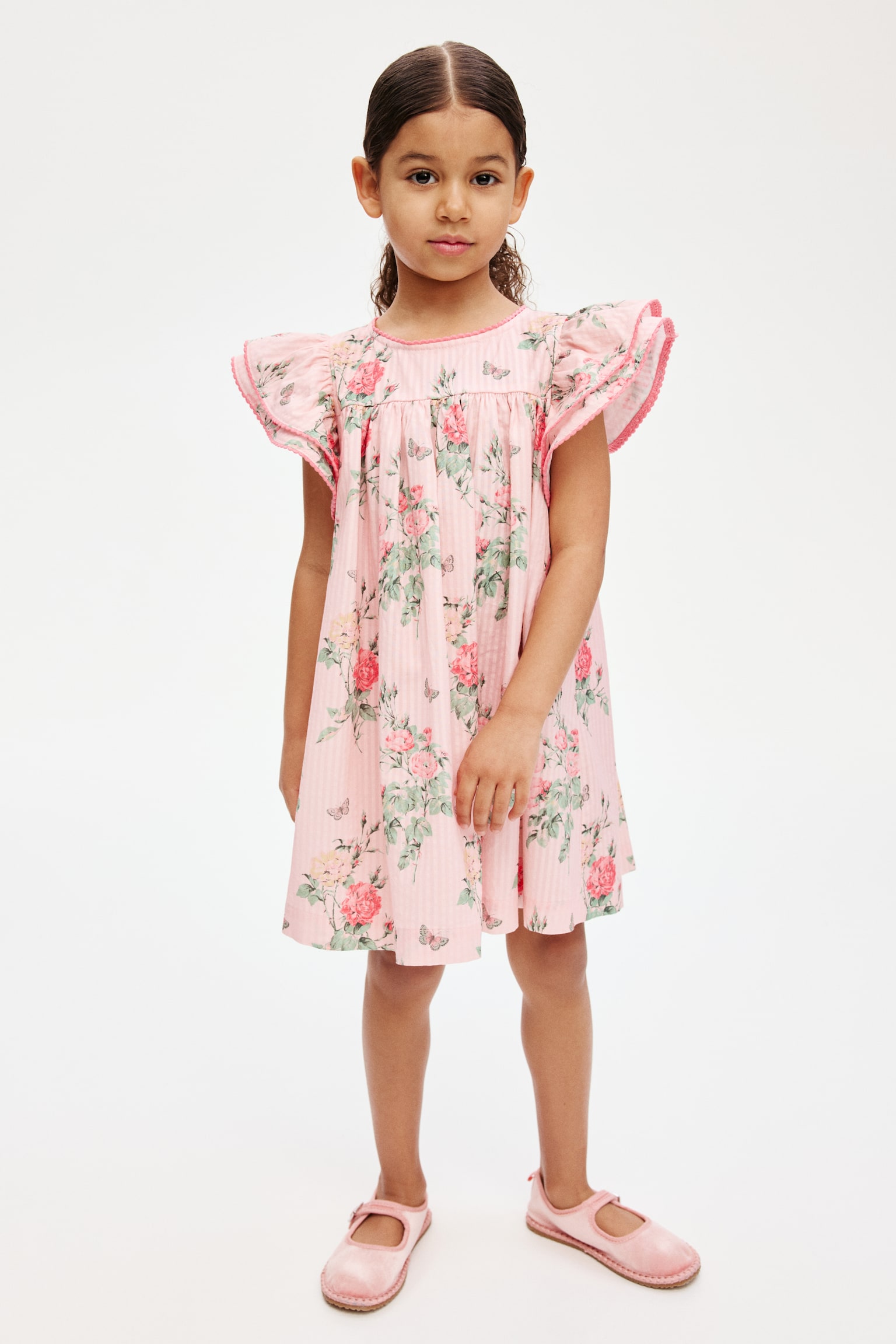 The empire-line dress, displayed from the front, showcases a pink cotton seersucker fabric adorned with a floral pattern of roses and butterflies. It features a round, picot-trimmed neckline and double-layered butterfly sleeves with picot trims. - 1