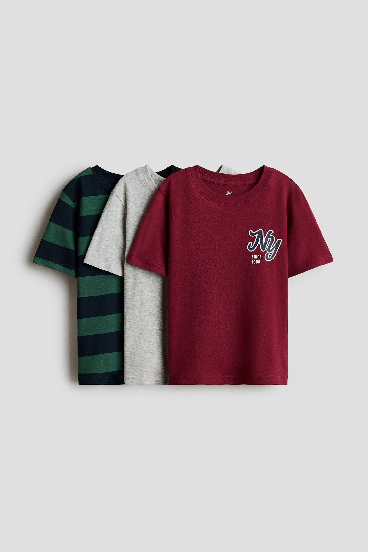 Three cotton jersey T-shirts are layered in a front-facing display. The front T-shirt is burgundy, featuring a rib-trimmed round neckline and a small "NY Since 1986" graphic on the chest, while the middle T-shirt is light grey and the back T-shirt features dark green and navy horizontal stripes.