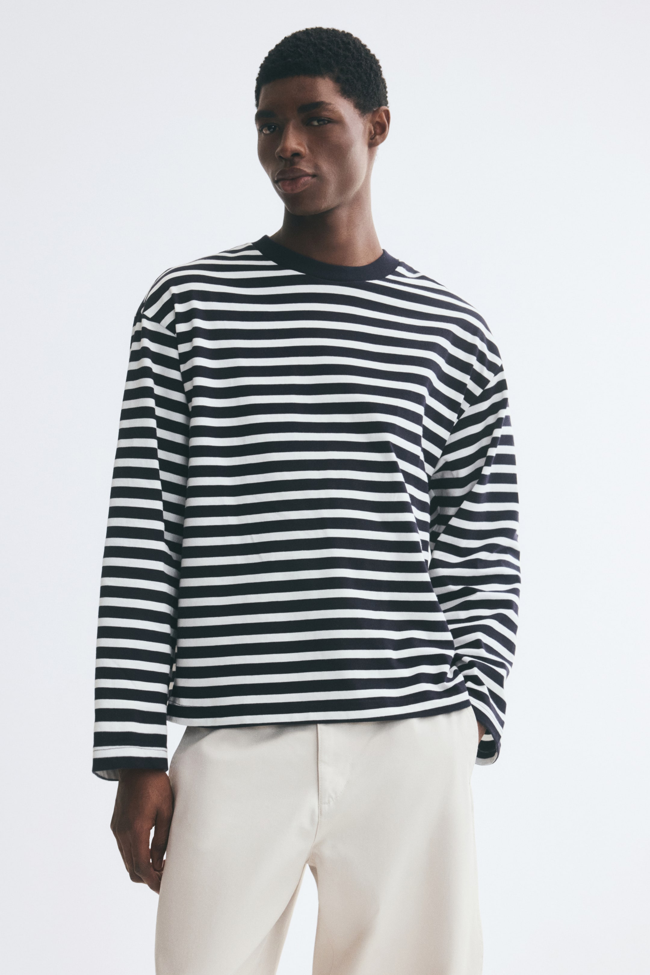 View larger image: The model is wearing a striped long-sleeve top with black and white horizontal bands, featuring an oversized fit, dropped shoulders, and a dark, rib-trimmed round neckline. The top is styled with light-colored trousers, with the model keeping one hand in a pocket.