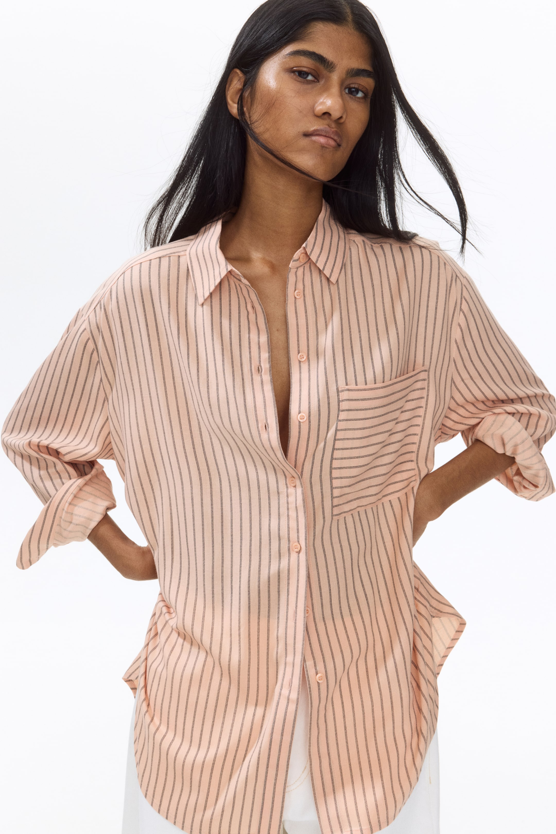 View larger image: The model is wearing an oversized, light peach shirt adorned with dark vertical stripes. This airy shirt is designed with a collar, a partially unbuttoned front, an open chest pocket, and relaxed, dropped shoulders. The long sleeves are rolled up, and the straight hem with rounded side vents is stylishly paired with white trousers. The model is casually placing her hands on her hips, adding a touch of elegance to the ensemble.