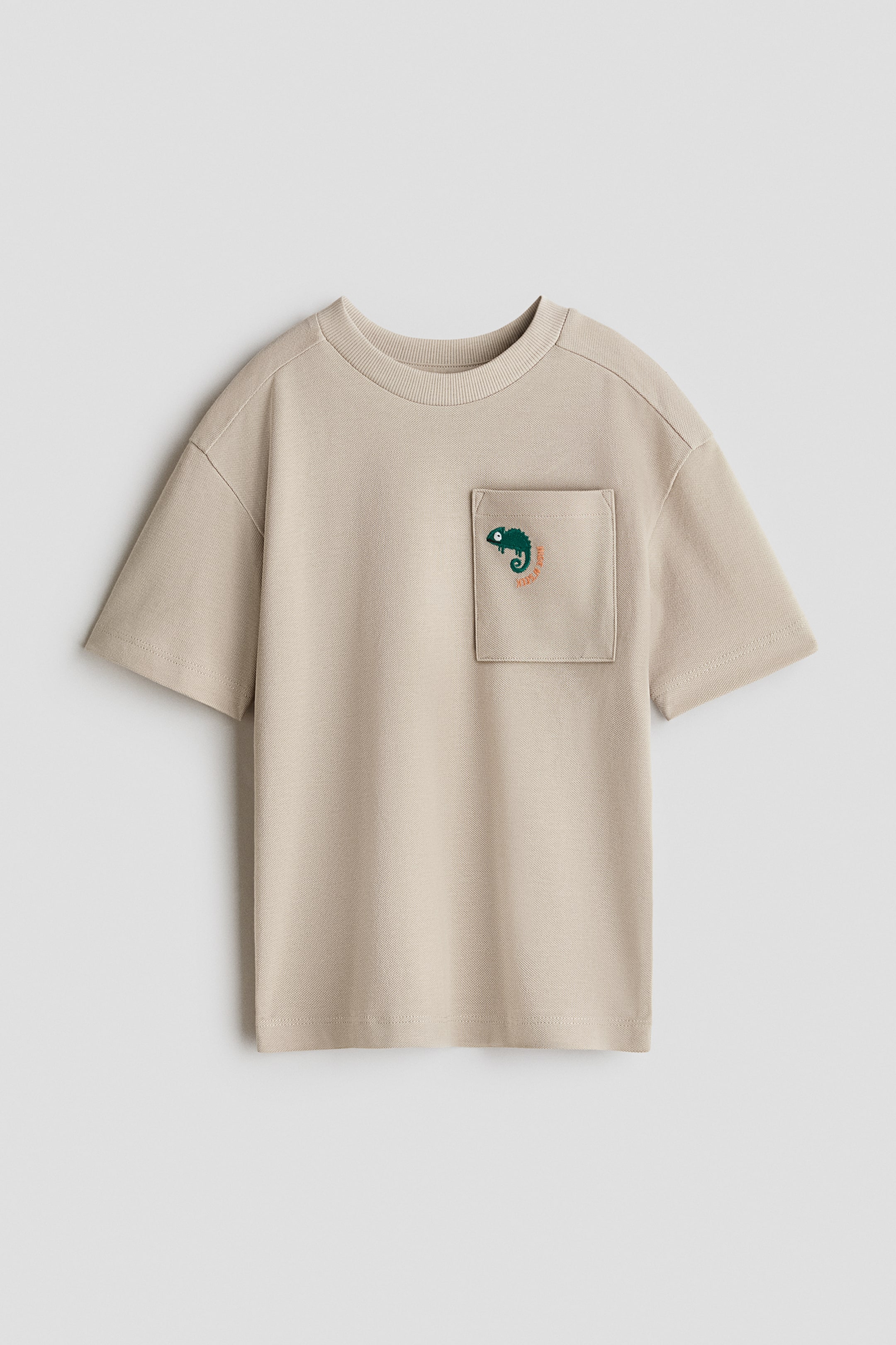 View larger image: A beige t-shirt featuring a ribbed crew neck and short sleeves. A square patch pocket on the left chest displays an embroidered green chameleon graphic.
