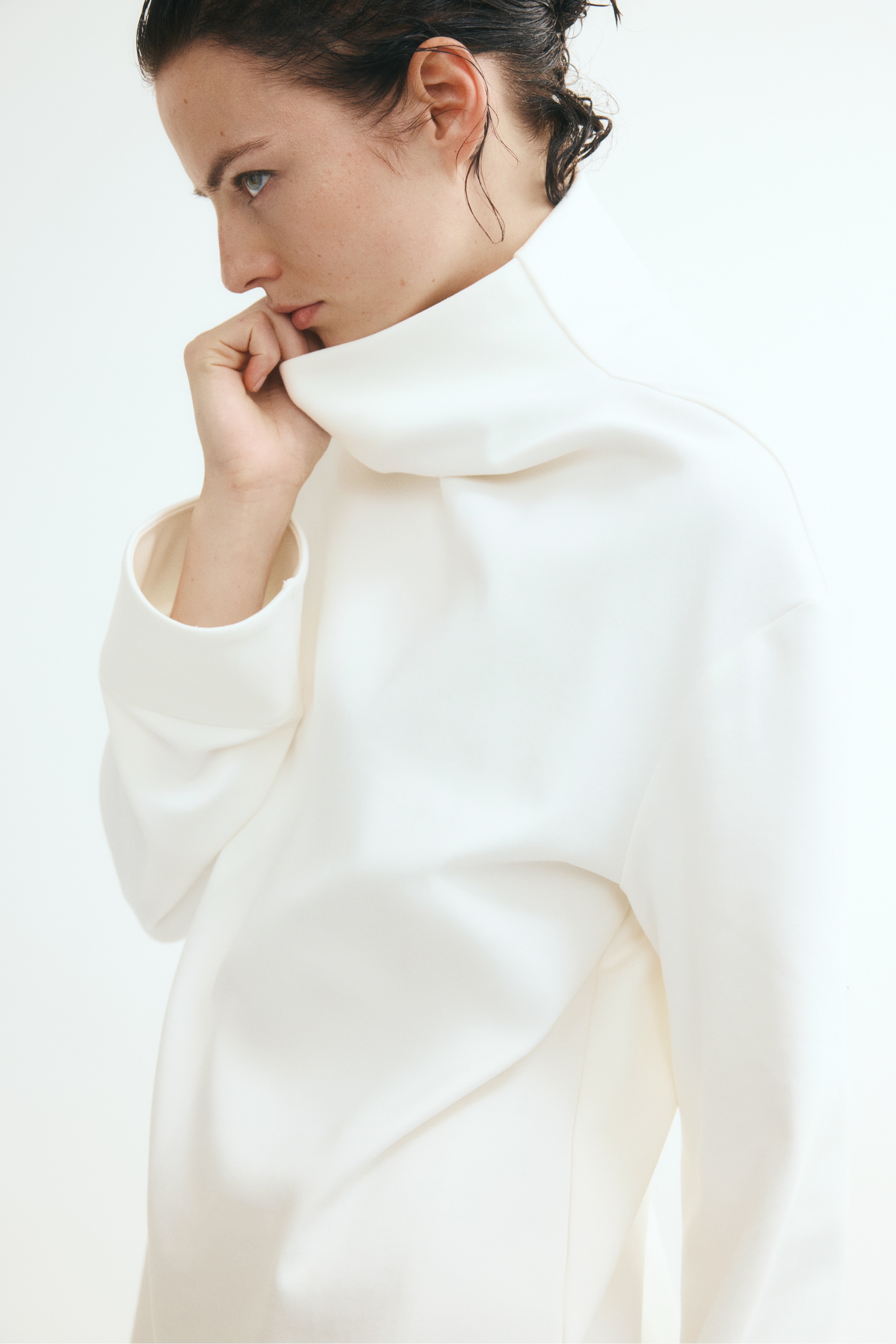 An off-white, high-neck top is shown from the back, featuring a relaxed fit with dropped shoulders and wide, long sleeves. The design showcases a clean, minimalist aesthetic with a straight hem and wide cuffs.
