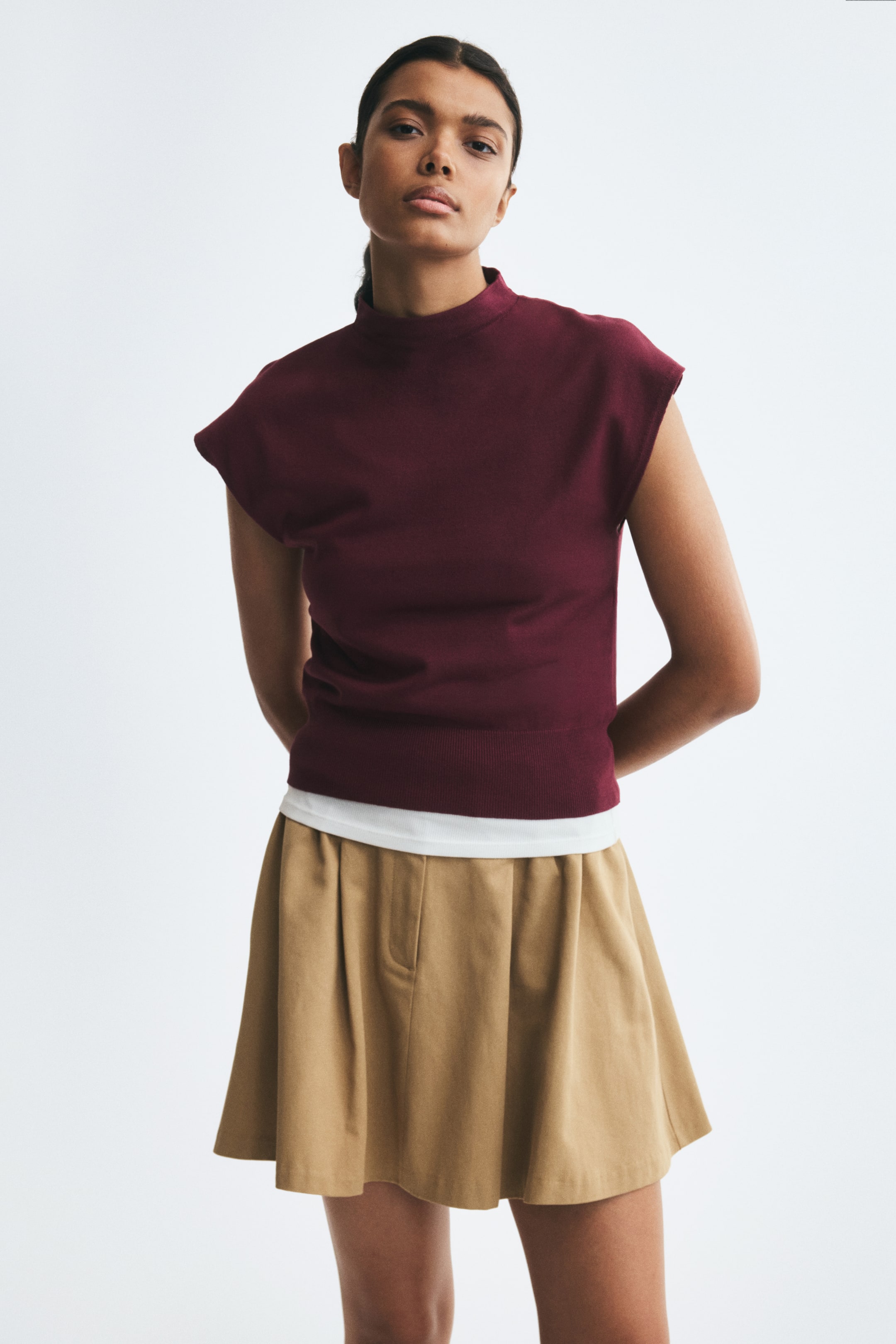 Ampliar la imagen: La modelo lleva un chaleco burdeos de punto fino y corto, con cuello alto y basta acanalada. El chaleco se lleva sobre una blusa blanca y se combina con una falda beige plisada.