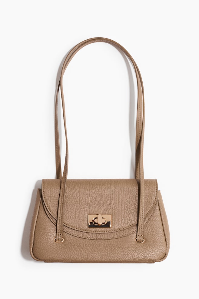 Un sac à main texturé couleur sable est présenté de face, avec un rabat arrondi fermé par un fermoir pivotant doré, et deux poignées fines et plates qui s'élèvent vers le haut à partir du dessus. - 1