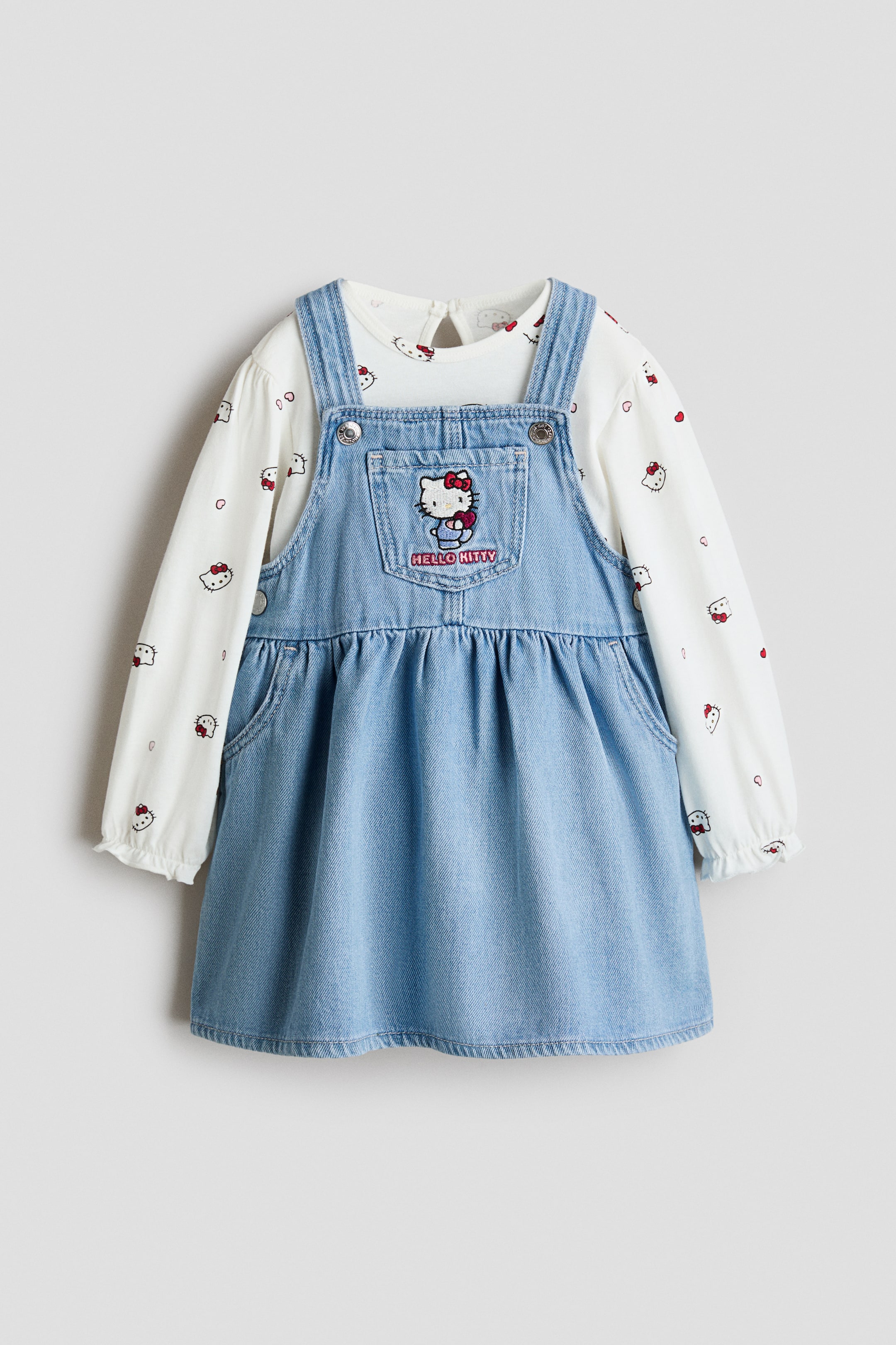 Xem ảnh lớn hơn: Bộ trang phục trẻ em gồm áo tay dài màu trắng mặc bên trong đầm yếm denim xanh nhạt, cả hai đều được chụp ở mặt trước. Áo có họa tiết in toàn bộ mặt Hello Kitty và trái tim đỏ, với bo thun ở cổ tay, trong khi đầm yếm denim nổi bật với dây đeo vai có thể điều chỉnh, túi bên hông và thiết kế Hello Kitty thêu trên túi yếm ở ngực.