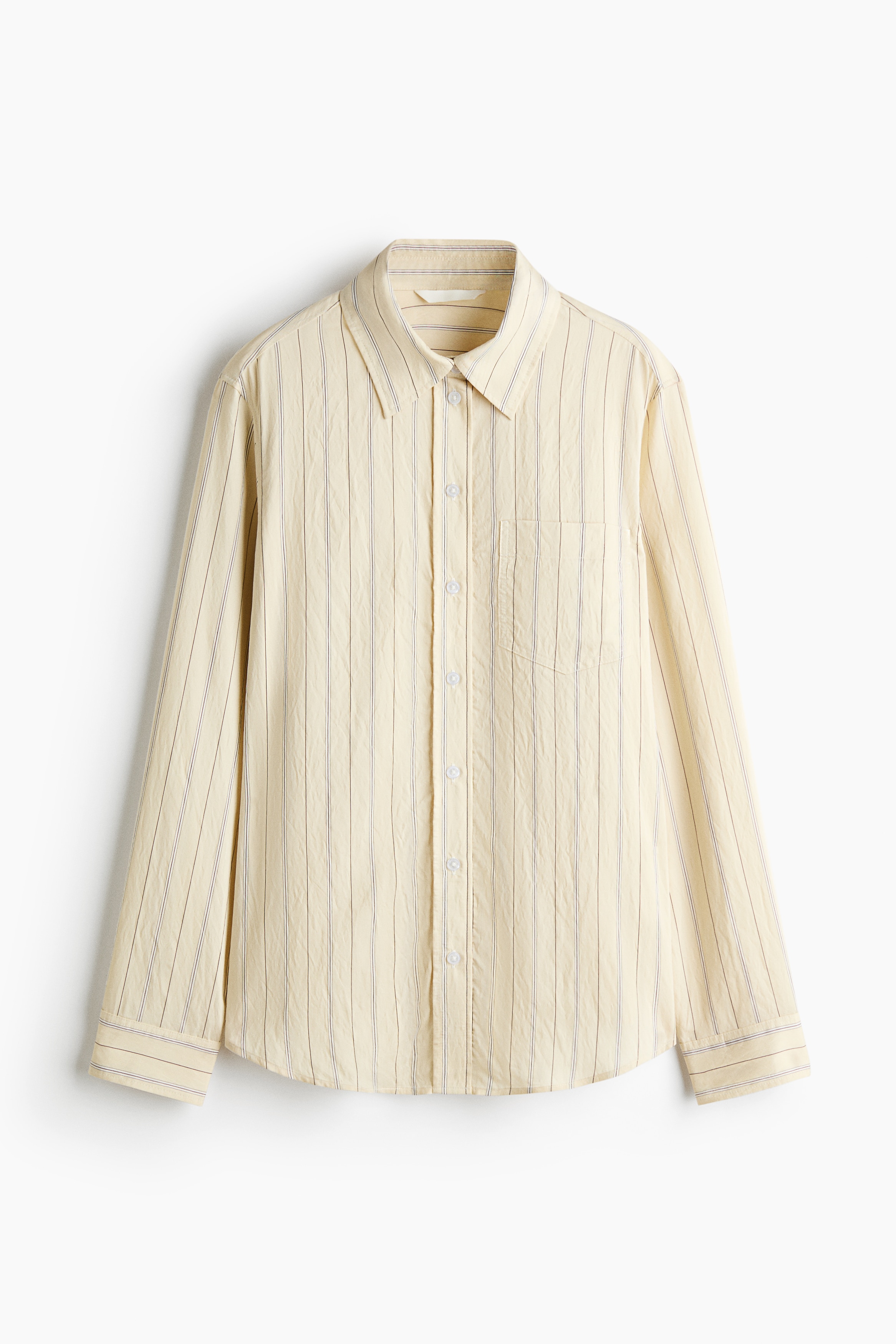 A light yellow, vertically striped button-down shirt is worn by the model, paired with wide-leg dark jeans and a white sweater draped over the shoulders. The long-sleeved shirt features a collar, a front button closure, and a rounded hem, with the model standing with one hand tucked into a pocket.