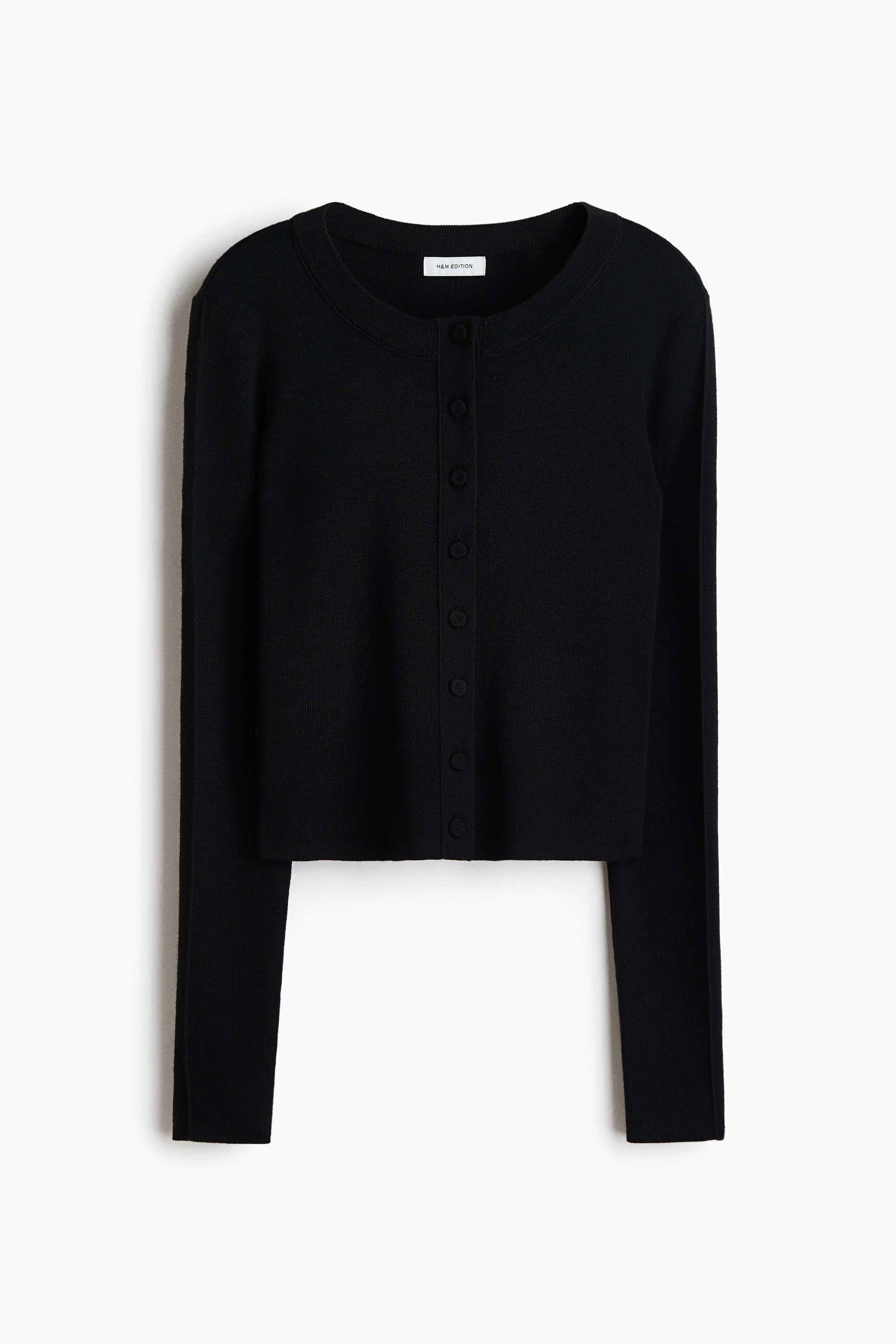 A short, fitted black cardigan in rib-knit wool with a round neckline, long sleeves and covered buttons is worn by the model. The cardigan is styled with matching slim-fit black trousers, and a black leather bag is carried under the modelâs arm.